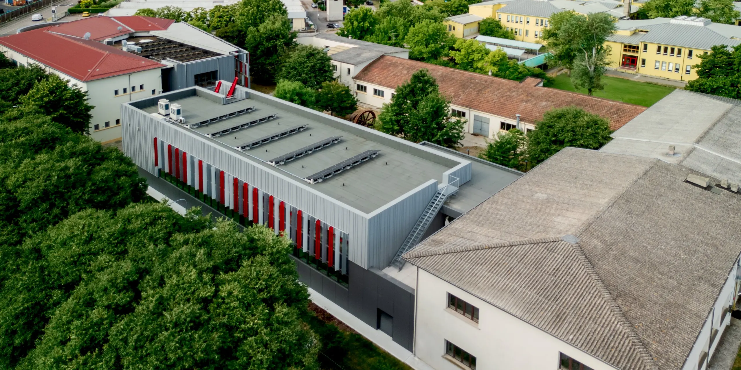 Vue aérienne impressionnante de la Scuola Arte e Mestieri F. Bertazzoni à Suzzara, nichée dans le paysage verdoyant de cette ville du nord de l'Italie. Le bâtiment moderne, avec sa façade continue en aluminium PREFA gris quartz et ses rayures rouges caractéristiques, se distingue avec assurance des édifices traditionnels environnants. La verticalité constante des éléments de façade PREFA et le toit plat percé de puits de lumière soulignent la clarté de la conception architecturale. Les produits en aluminium PREFA garantissent une valeur durable, une grande robustesse et une esthétique toujours séduisante.