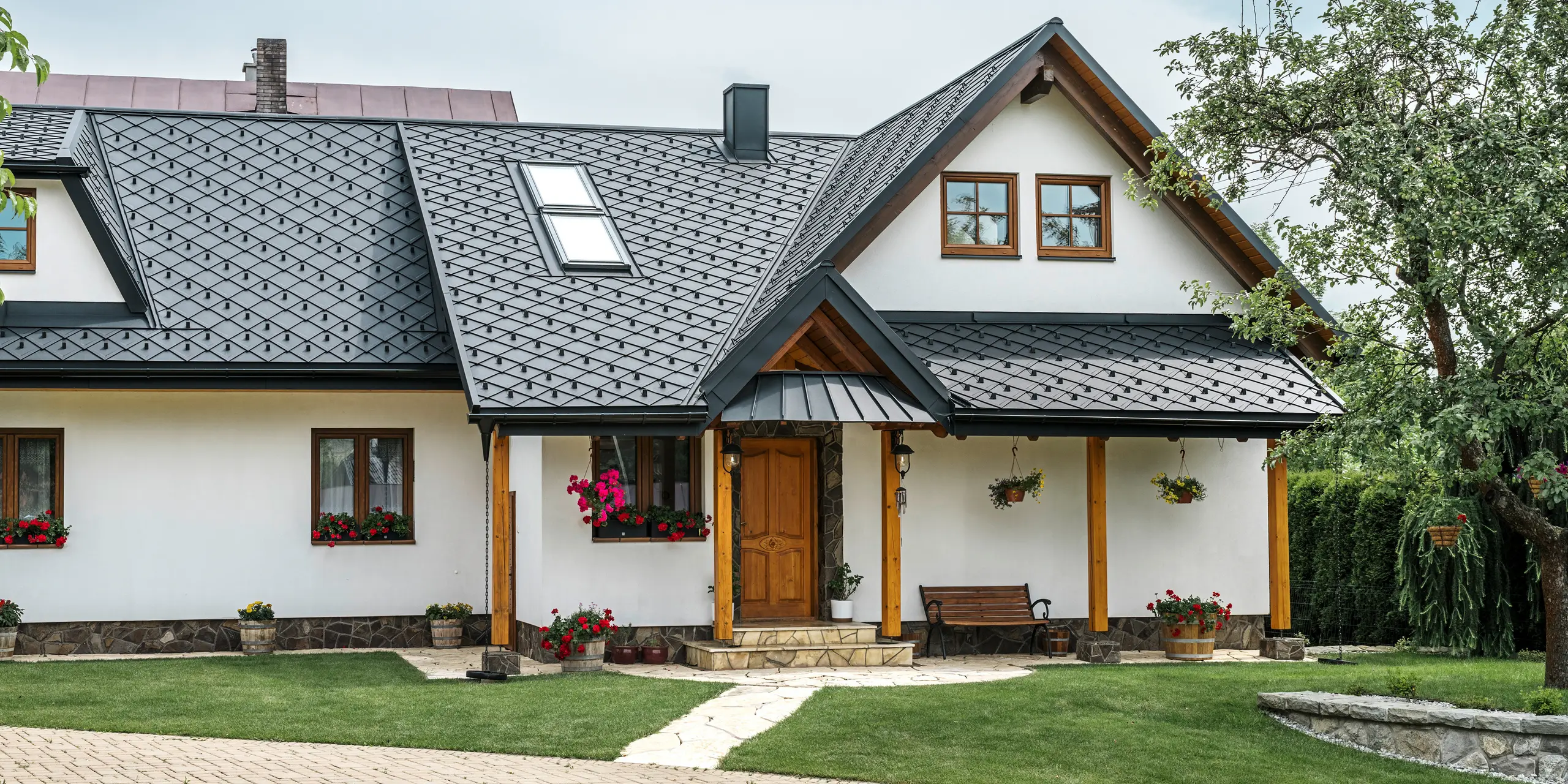 Zone d'entrée d'une maison familiale traditionnelle à Stara Bystrica, Slovaquie, avec un toit en aluminium composé de losanges PREFA 29 × 29 en P.10 noir. Le toit en aluminium de haute qualité au look élégant comprend des fenêtres de toit inclinées et une véranda avec auvent recouvert de PREFALZ. Les produits en aluminium à la fois élégants et robustes confèrent à la maison une apparence contemporaine et accueillante. Le jardin bien entretenu et les compositions florales soulignent le bon entretien du bâtiment.