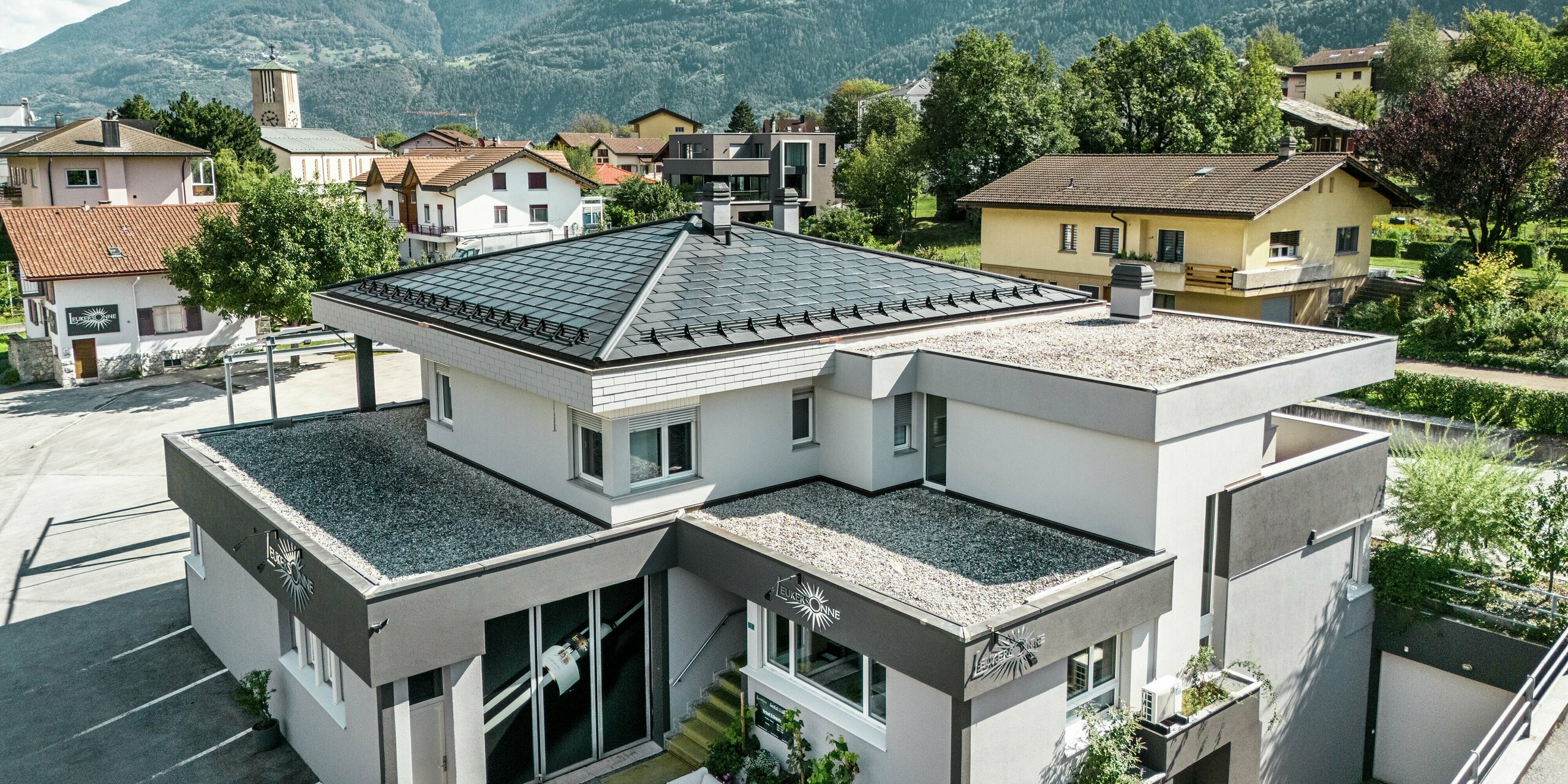 L'élégant toit en aluminium PREFA confère à la cave Leukersonne à Susten (CH) un aspect moderne et offre une protection optimale contre les intempéries. Les tuiles solaires PREFA garantissent une production d'énergie durable sans compromettre l'apparence du toit de la tente. Le paysage du toit est complété par des R.16 PREFA. Le premier étage et la partie restante du deuxième étage sont recouverts d'une surface en gravier praticable.