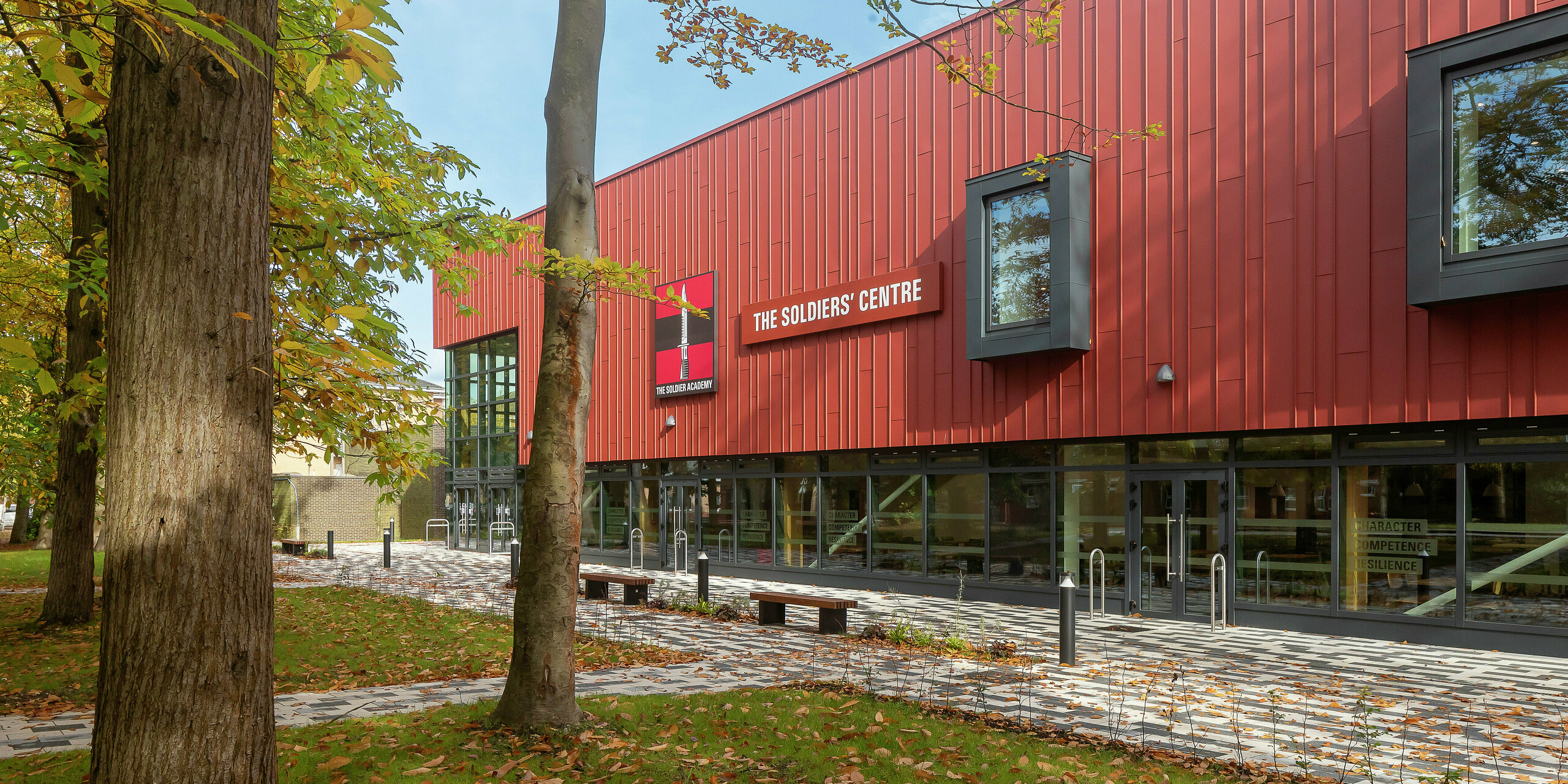 Vue détaillée des éléments de façade modernes du Soldiers' Centre de Pirbright : Le point central est le revêtement à joint debout précisément installé en PREFALZ en P.10 rouge oxyde, qui, en combinaison avec les cadres de fenêtres marquants en P.10 anthracite, met un accent visuel. Les lignes fines et les effets d'ombre soulignent la finition de haute qualité et le design innovant des produits en aluminium PREFA - un témoignage d'une architecture durable et contemporaine.