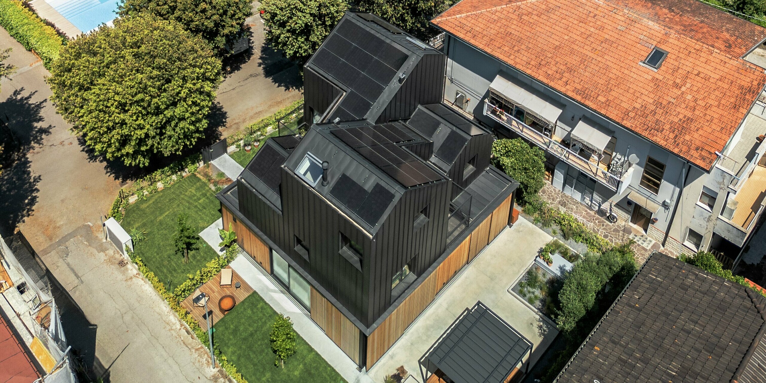 Vue aérienne d'une maison jumelée moderne à Cattolica : façade et toiture en aluminium PREFALZ gris noir, associées à des lattes de bois. Le bâtiment se distingue par ses lignes épurées et la qualité de ses matériaux PREFA, contrastant avec les maisons traditionnelles aux toits de tuiles rouges et le cadre méditerranéen.