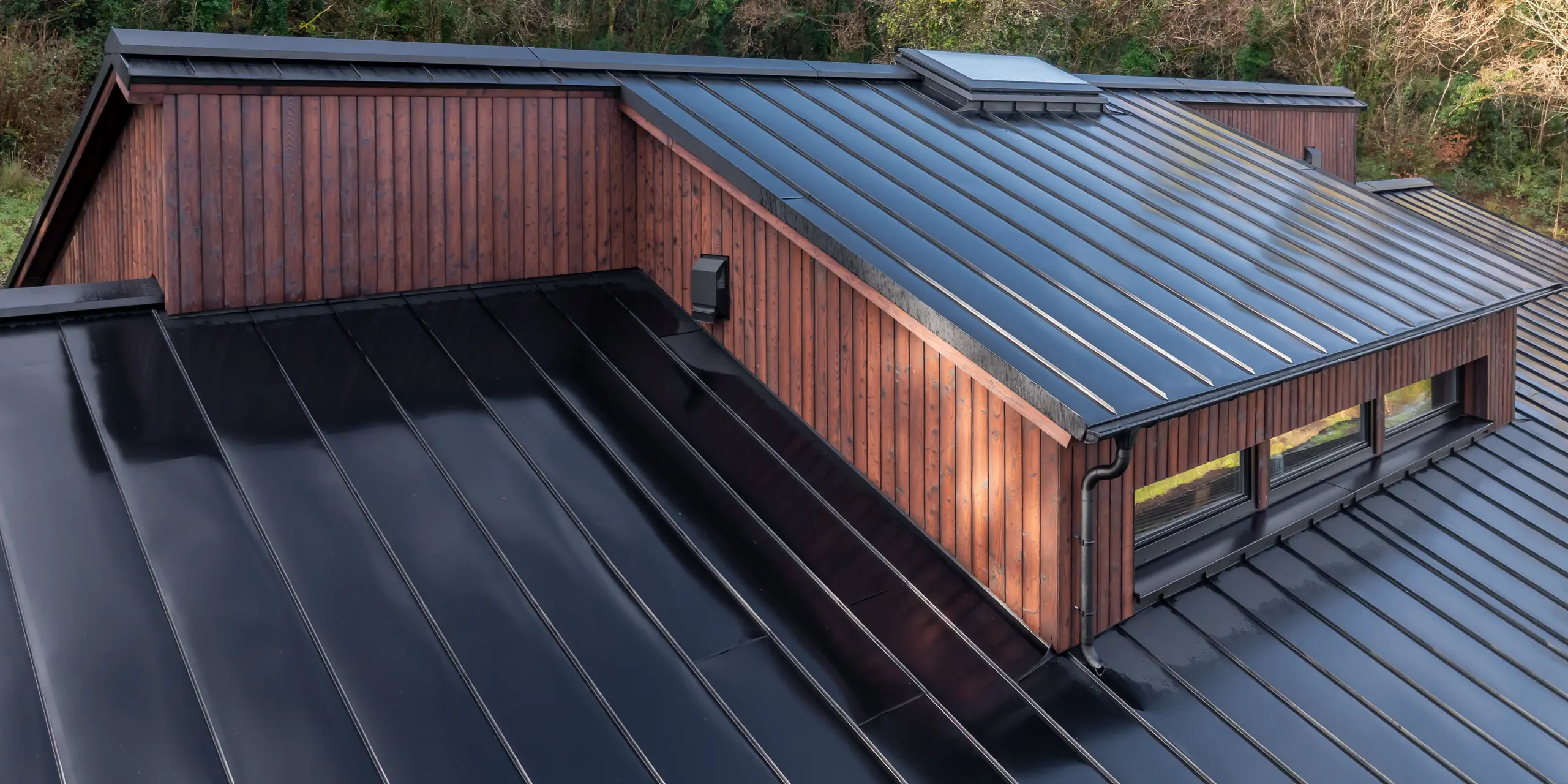 Vue oblique de la toiture de la Maison Holywell à Sligo : les lignes épurées de la toiture en aluminium PREFALZ P.10 Noir se déploient avec dynamisme sur plusieurs niveaux, attirant le regard vers la lucarne à l’exécution précise. Les joints debout réguliers reflètent la lumière et soulignent la qualité de la réalisation et la précision des assemblages. Associée à la façade en bois, la toiture crée une image architecturale forte, renforcée par la durabilité, la résistance à la corrosion et le faible entretien des produits en aluminium PREFA.