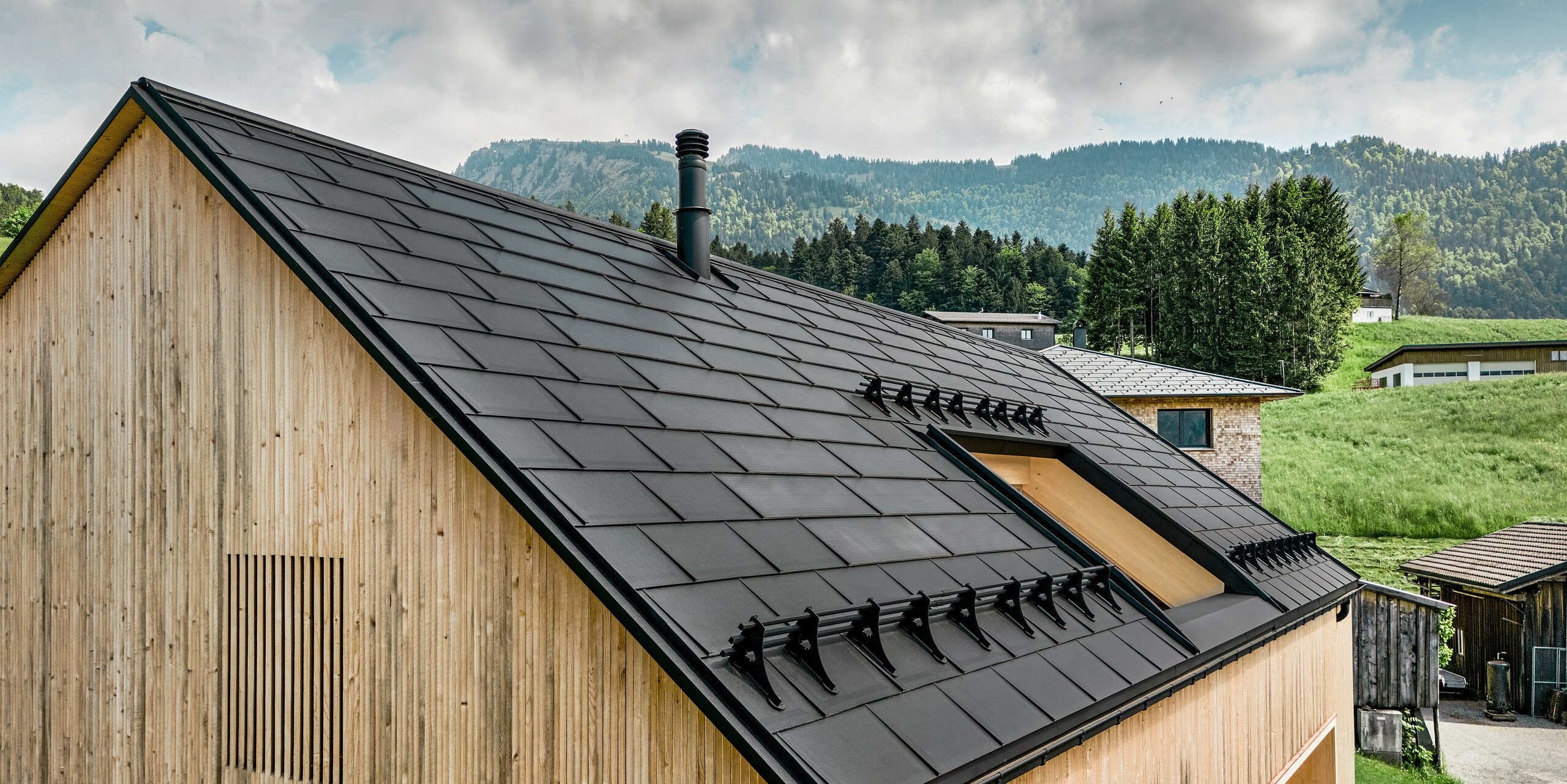 Vue détaillée d'une maison écologique à Egg, équipée de tuiles solaires PREFA efficaces, qui se distinguent parmi les R.16 PREFA de haute qualité. Le bois naturel de la façade crée un contraste chaleureux avec le style moderne du toit en aluminium noir, qui allie efficacité énergétique et esthétique élégante. Le toit, surmonté d'une fine cheminée, reflète une construction durable ancrée dans un cadre rural de collines et de verdure luxuriante.