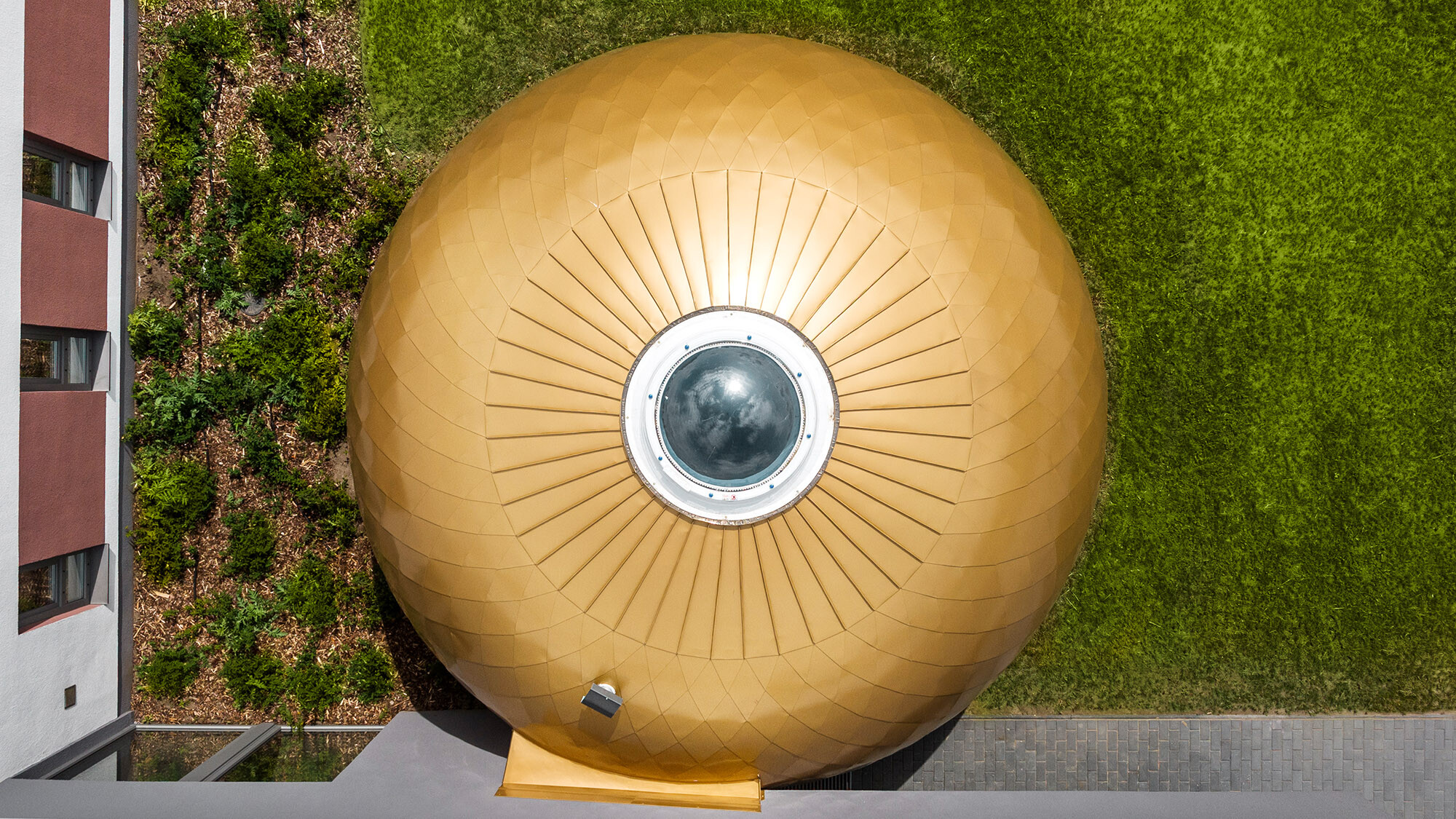L’une des pièces rondes avec puits de lumière vue d’en haut ; en bas et à gauche de l’image elle est encadrée par la façade en crépi, en haut et à droite de l’image s’étend la pelouse.