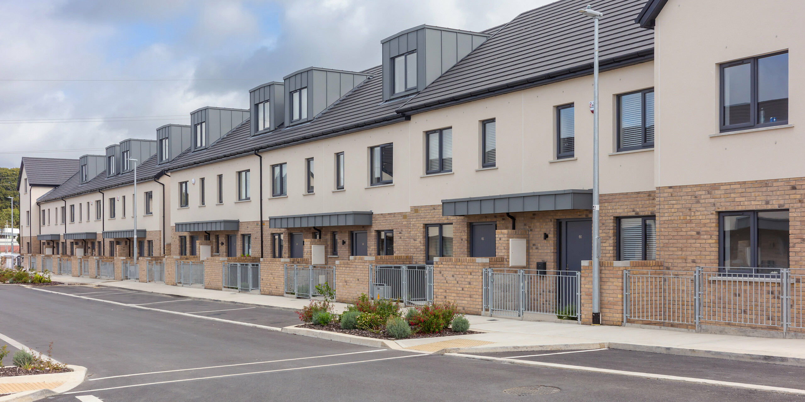 La photographie panoramique présente un ensemble de maisons mitoyennes modernes à Churchfields, dont l'aspect uniforme est fortement influencé par les lucarnes revêtues de profilés PREFALZ P.10 gris souris. Les lignes épurées des joints debout de l'aluminium contrastent élégamment avec le matériau de toiture sombre, conférant à la longue rangée de maisons une structure moderne et rythmée. Les éléments PREFA, robustes, sont inaltérables, résistants aux intempéries et ne nécessitent pratiquement aucun entretien – une solution idéale pour les grands projets résidentiels. Les jardins de façade soignés et la vue paisible sur la rue soulignent l'harmonie de l'ensemble.