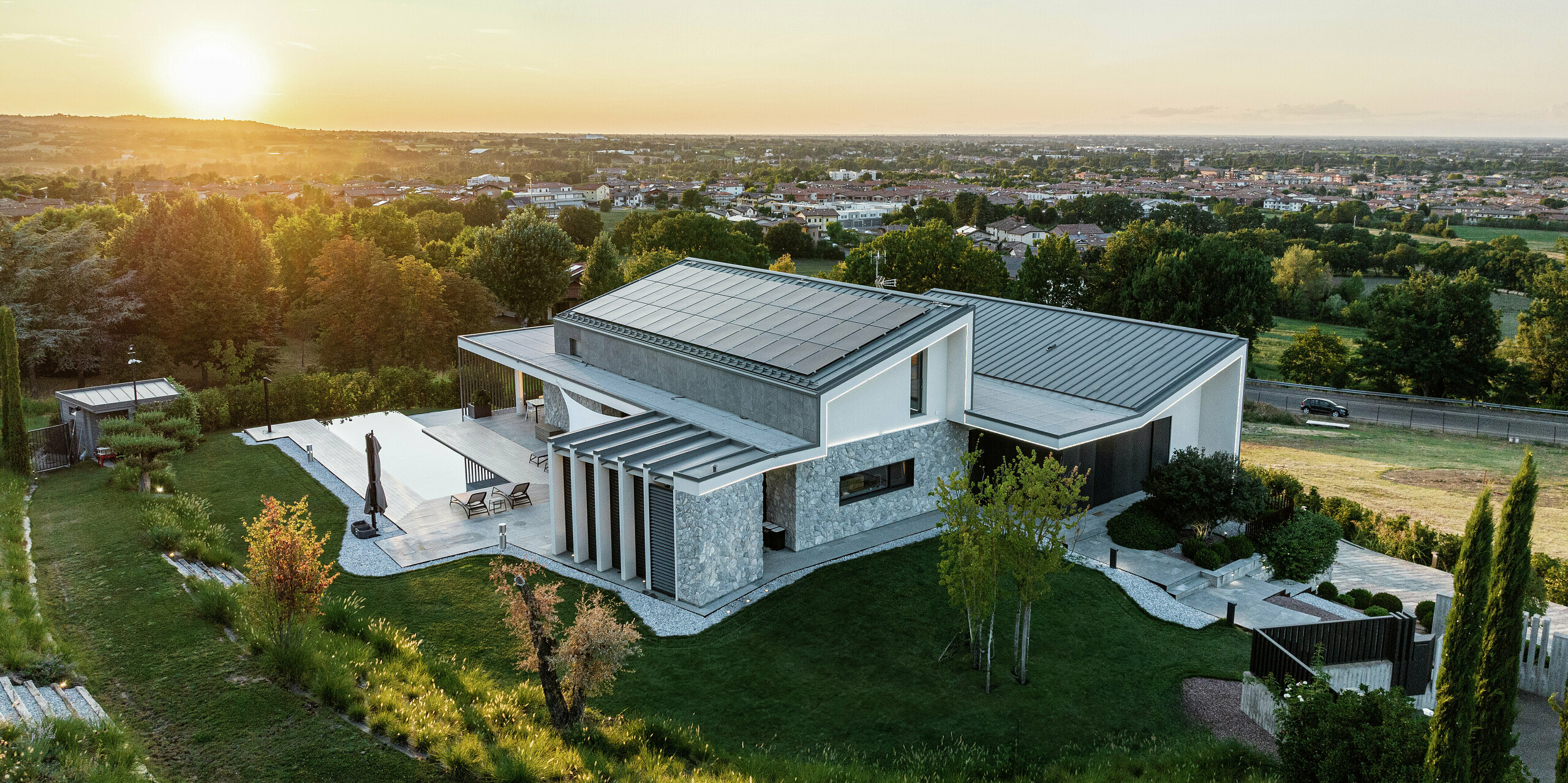 La maison unifamiliale à Scandiano, en Italie, dans la chaude lumière du soir. Les deux toits en pente avec PREFALZ en P.10 anthracite soulignent l'architecture exclusive et offrent une durabilité et une résistance maximales. La terrasse spacieuse avec piscine, le paysage de jardin harmonieux et la conception moderne de la façade complètent le tableau d'ensemble luxueux. Le système photovoltaïque à grande échelle assure une production d’énergie respectueuse de l’environnement à partir de l’énergie solaire.