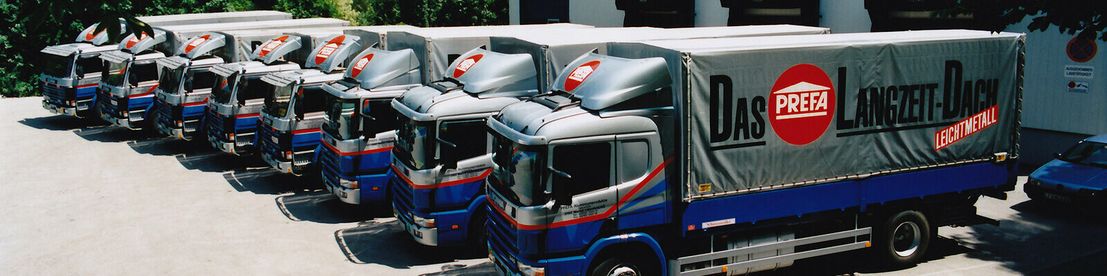 Huit anciens camions PREFA en bleu et argent avec logo PREFA alignés 