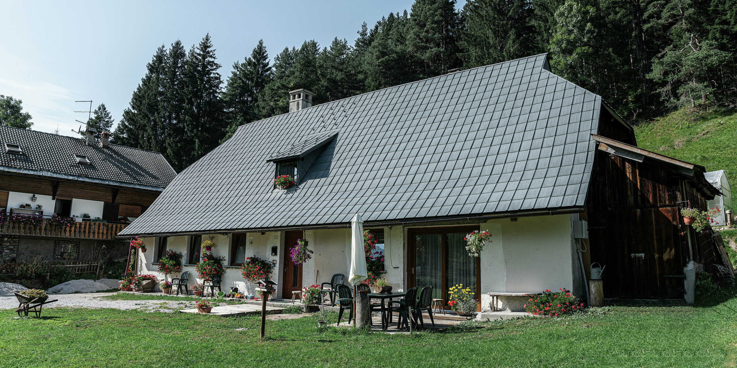 Une charmante maison unifamiliale à Tarvisio, en Italie, est recouverte de tuiles PREFA durables depuis 1972 : le grand toit en croupe avec son revêtement en aluminium durable protège de manière fiable la maison des intempéries alpines depuis des décennies. Des jardinières fleuries ornent la façade rustique et une terrasse chaleureuse avec mobilier de jardin vous invite à vous attarder. En arrière-plan, des conifères denses et verts créent une atmosphère idyllique et intègrent parfaitement la maison dans l'environnement naturel.