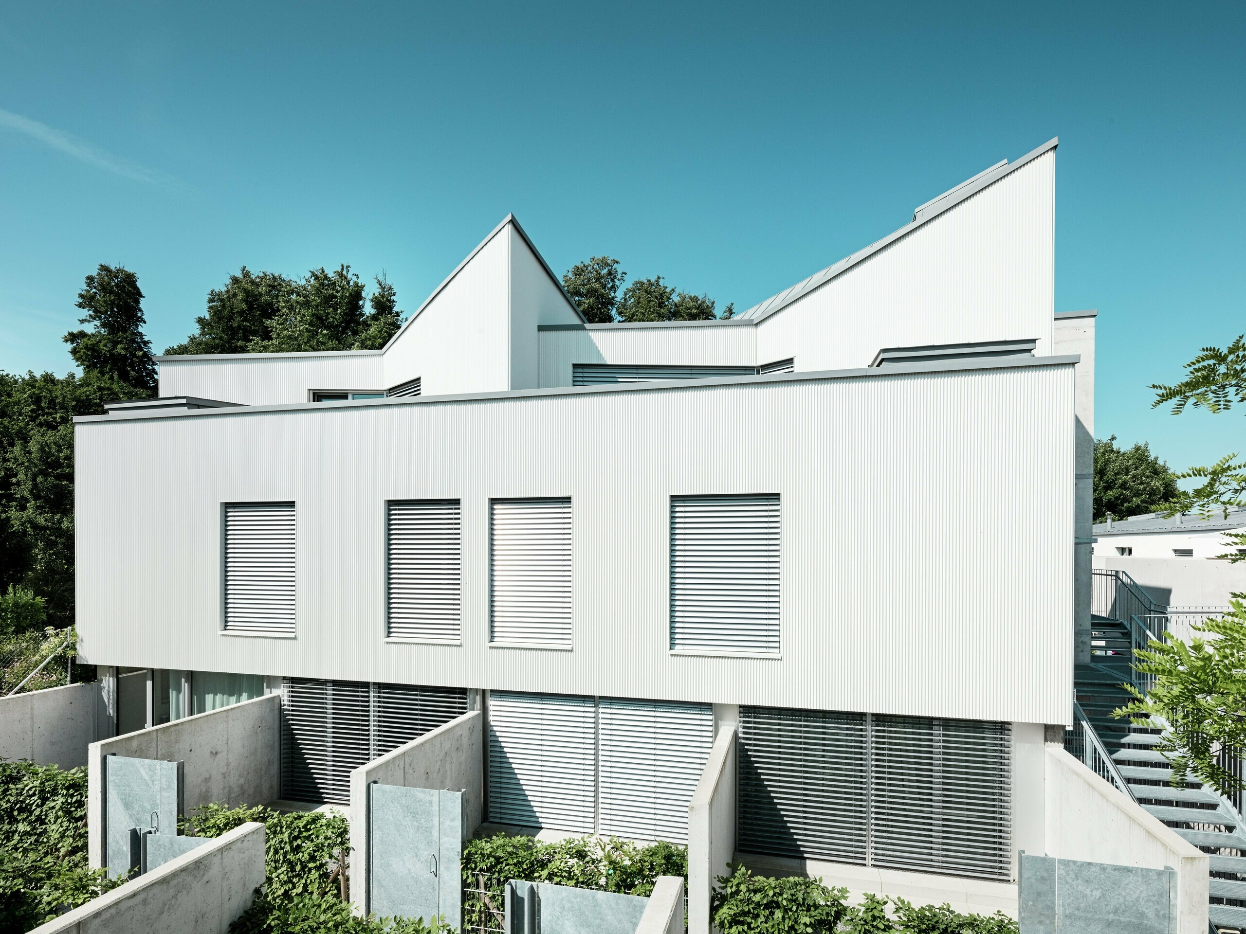 Les Tiny Homes contemporaines situées à Zollikerberg, en Suisse, séduisent par leurs lignes épurées et leur façade moderne en aluminium PREFA avec le profil extrudé « Triangle ». Les profils posés verticalement, aux courbes douces, créent une façade à la fois marquante et élégante, animée par les reflets de la lumière. La surface blanche apporte une sensation de légèreté et souligne l’architecture géométrique. La façade rapportée et ventilée protège durablement le bâtiment contre les intempéries et améliore le confort intérieur. Elle est facile d’entretien et résistante à la corrosion.