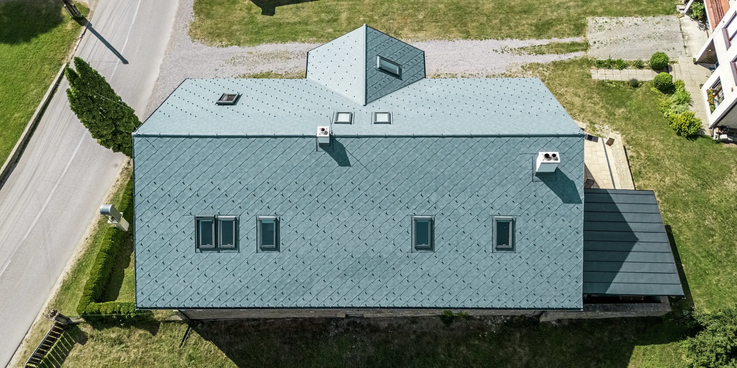 L'image présente une vue aérienne impressionnante du toit d'une maison d'artisan à Klin, en Slovaquie. La disposition symétrique des losanges PREFA 44 x 44, couleur Gris Pierre P.10, souligne l'élégance architecturale de la maison. Les nombreuses fenêtres de toit s'intègrent harmonieusement à la charpente et créent une atmosphère lumineuse et accueillante à l'intérieur. Outre son attrait esthétique, la toiture en aluminium offre des avantages pratiques : exceptionnellement durable, résistante à la corrosion et légère, elle constitue le choix idéal pour la rénovation d'un bâtiment historique. Cette solution de toiture allie une esthétique parfaite à une fonctionnalité optimale et une résistance aux intempéries.