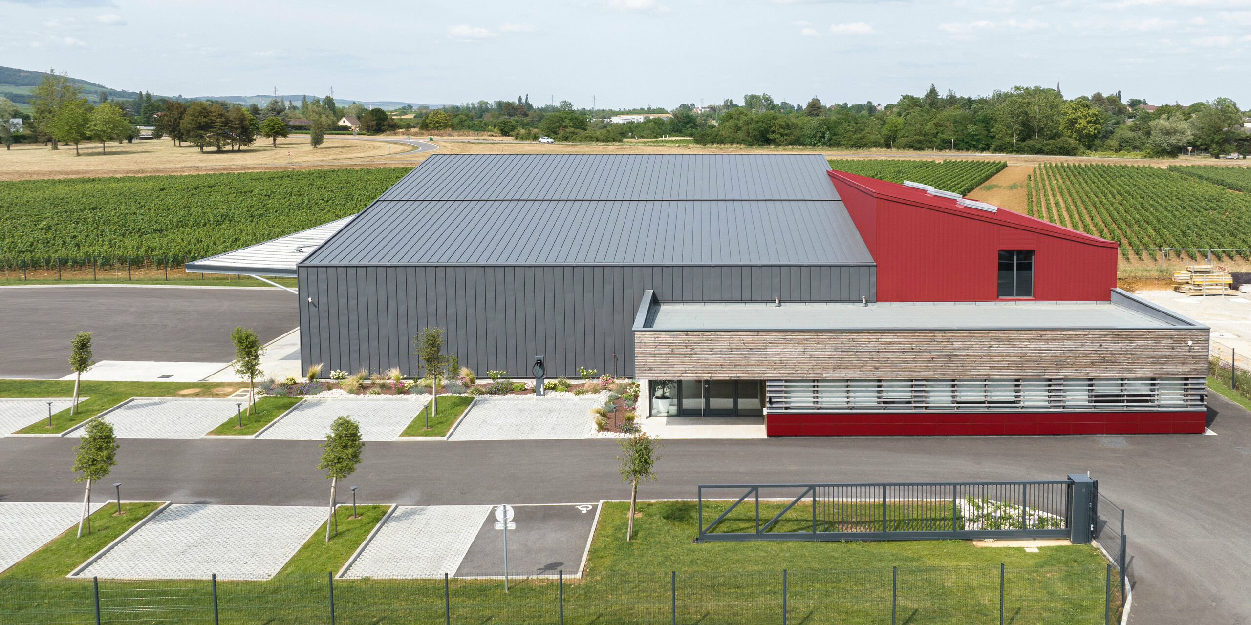 Un grand bâtiment industriel avec PREFALZ en gris sombre sur le toit et la façade. Pour accentuer la couleur, certaines parties du bâtiment ont été revêtues de revêtements PREFALZ ou PREFA en P.10 rouge oxyde. Le bâtiment se dresse face à un paysage avec des champs en arrière-plan. Sur le bord avant de l'image, vous pouvez voir des places de parking avec de jeunes arbres et une plantation soignée.