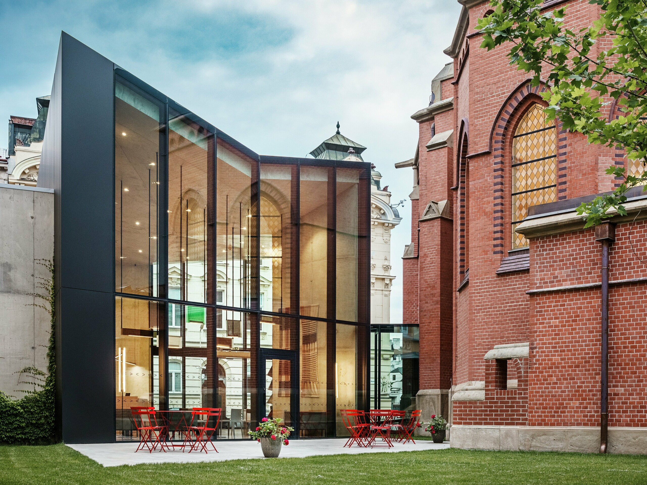 L'image révèle le fascinant nouveau bâtiment d'Olomouc, qui relie l'église rouge historique à une bibliothèque moderne. La façade noire, réalisée en panneaux composites en aluminium PREFABOND, contraste fortement avec la chaleur des briques rouges de l'église. De larges surfaces vitrées offrent une vue dégagée sur l'intérieur, où règne une atmosphère moderne et lumineuse. Sur la terrasse, tables et chaises rouges invitent à la détente et créent un lien harmonieux entre la structure historique et l'architecture contemporaine. Cette élégante intégration de l'ancien et du nouveau illustre les atouts des panneaux PREFABOND : outre leur esthétique moderne, ils offrent une durabilité et une fonctionnalité exceptionnelles.