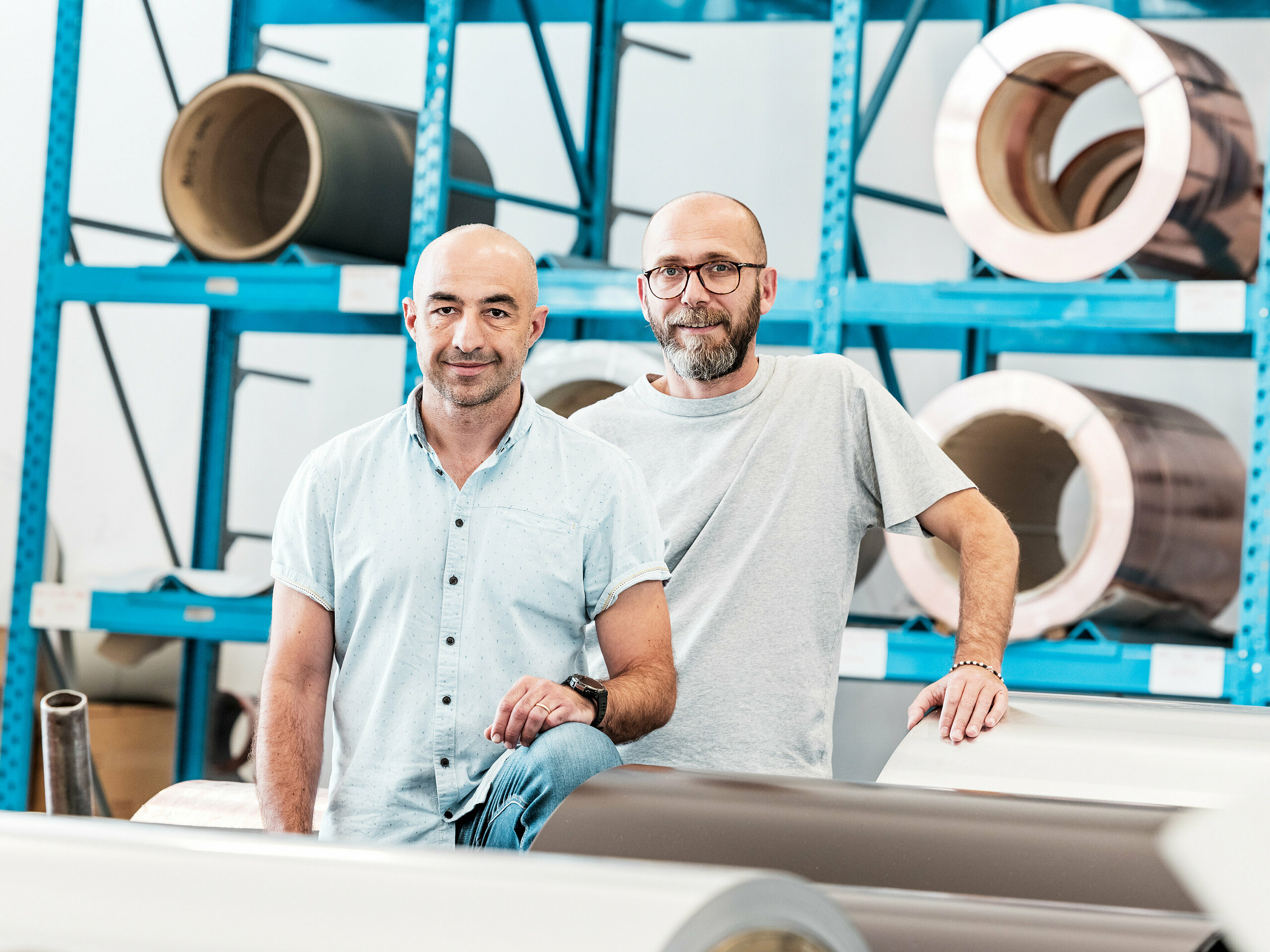 Deux artisans dans l'entrepôt d'aluminium devant des bobines métalliques enroulées – un environnement de travail moderne pour les systèmes de toiture et de façade en aluminium.