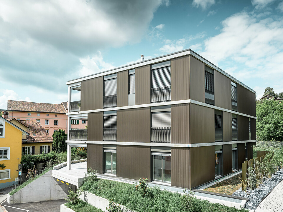 Die markante Aluminiumfassade dieses Mehrfamilienhauses in Schaffhausen ist mit dem eleganten PREFA Zackenprofil in der Farbe Colinal ausgestattet. Durch die vertikale Anordnung der stranggepressten Aluminiumprofile wirkt das Gebäude optisch kompakter und die Fassadenstruktur erhält eine harmonische Balance. Das edle Design fügt sich geschmackvoll in die Umgebung ein und verleiht dem Gebäude eine moderne Eleganz, die sowohl optisch ansprechend als auch funktional ist. Die Perspektive zeigt das Mehrfamilienhaus von der Vorderseite mit der Garageneinfahrt. Im Hintergrund sind Bäume, weitere Gebäude und ein Kirchturm zu erkennen.