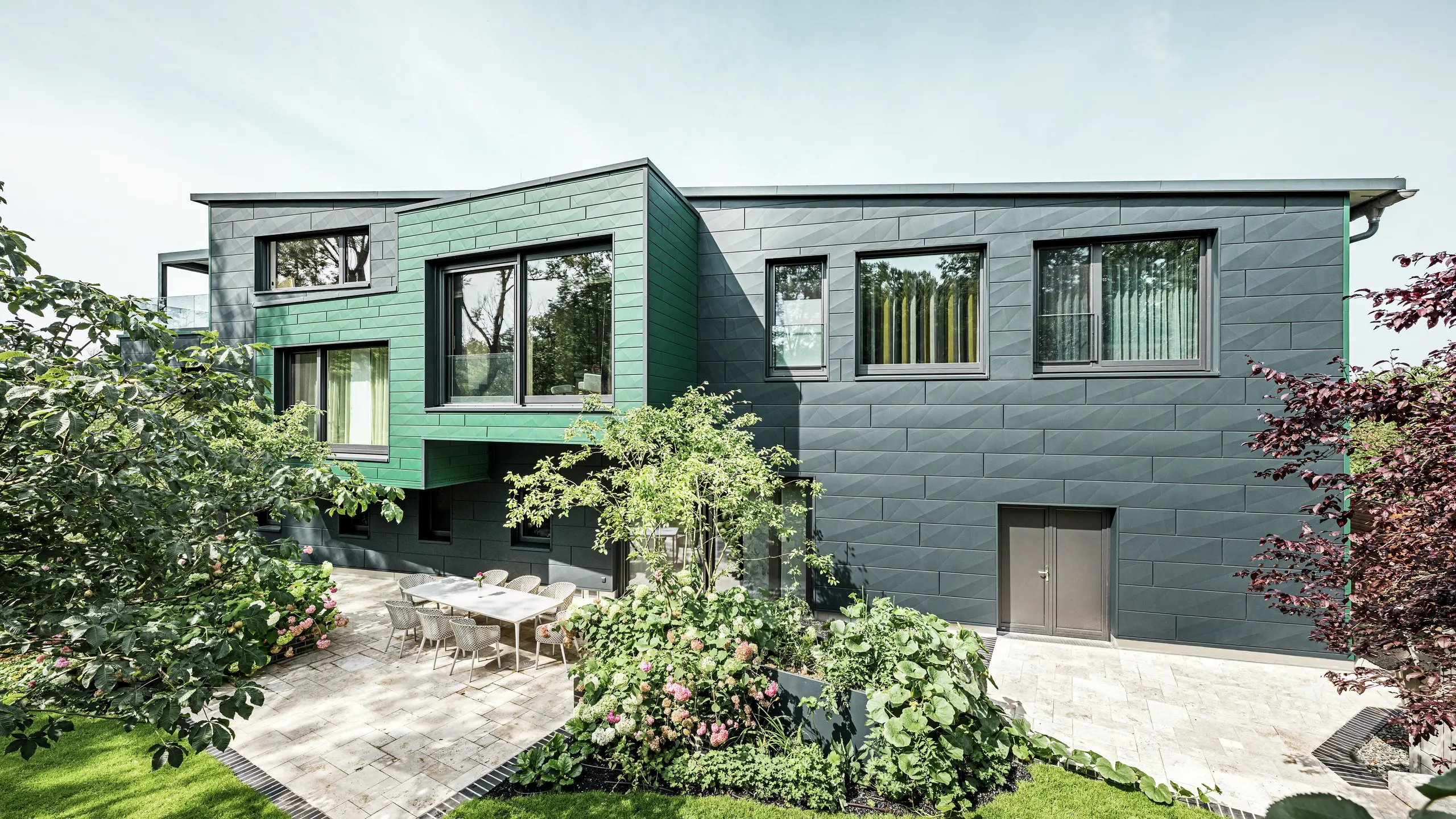 Cette maison individuelle à Türkheim impressionne par sa façade exceptionnelle, revêtue de panneaux PREFA Siding X anthracite et vert mousse. La pureté des lignes et le choix audacieux des couleurs des panneaux d'aluminium confèrent au bâtiment une allure moderne et élégante. Le vaste jardin, agrémenté d'une terrasse raffinée et d'une table à manger entourée de chaises contemporaines, est mis en valeur. Des massifs de fleurs colorés et des plantes luxuriantes offrent un contraste verdoyant et dynamique à la façade robuste. Durable et nécessitant peu d'entretien, la façade en aluminium protège le bâtiment et lui assure une beauté intemporelle.