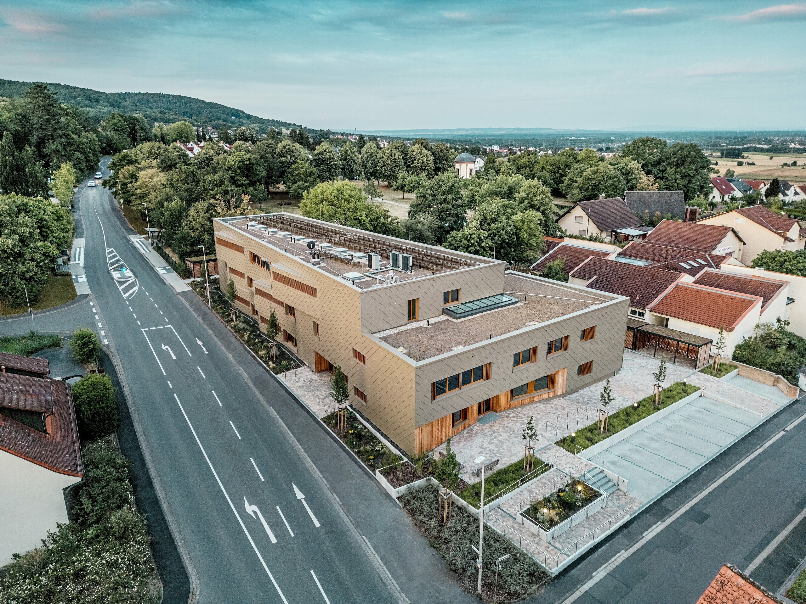 La conception globale impressionnante de la crèche municipale d'Alzenau est clairement visible sur la photographie aérienne. Ce bâtiment hybride aux lignes angulaires s'intègre harmonieusement dans un quartier résidentiel paisible, en bordure de la forêt de Spessart. La façade, composée de losanges de façade 44x44 PREFA couleur bronze, confère au bâtiment une allure remarquable et de grande qualité, et souligne son architecture moderne. Toitures végétalisées, vastes espaces extérieurs et circulation fluide témoignent de la conception durable du projet. L'association de matériaux durables comme l'aluminium, le bois et le béton recyclé répond aux normes écologiques les plus exigeantes. Cette perspective met en lumière l'intégration urbaine réfléchie et la multifonctionnalité du bâtiment – ​​un projet novateur en matière d'éducation, de logement et de développement durable.