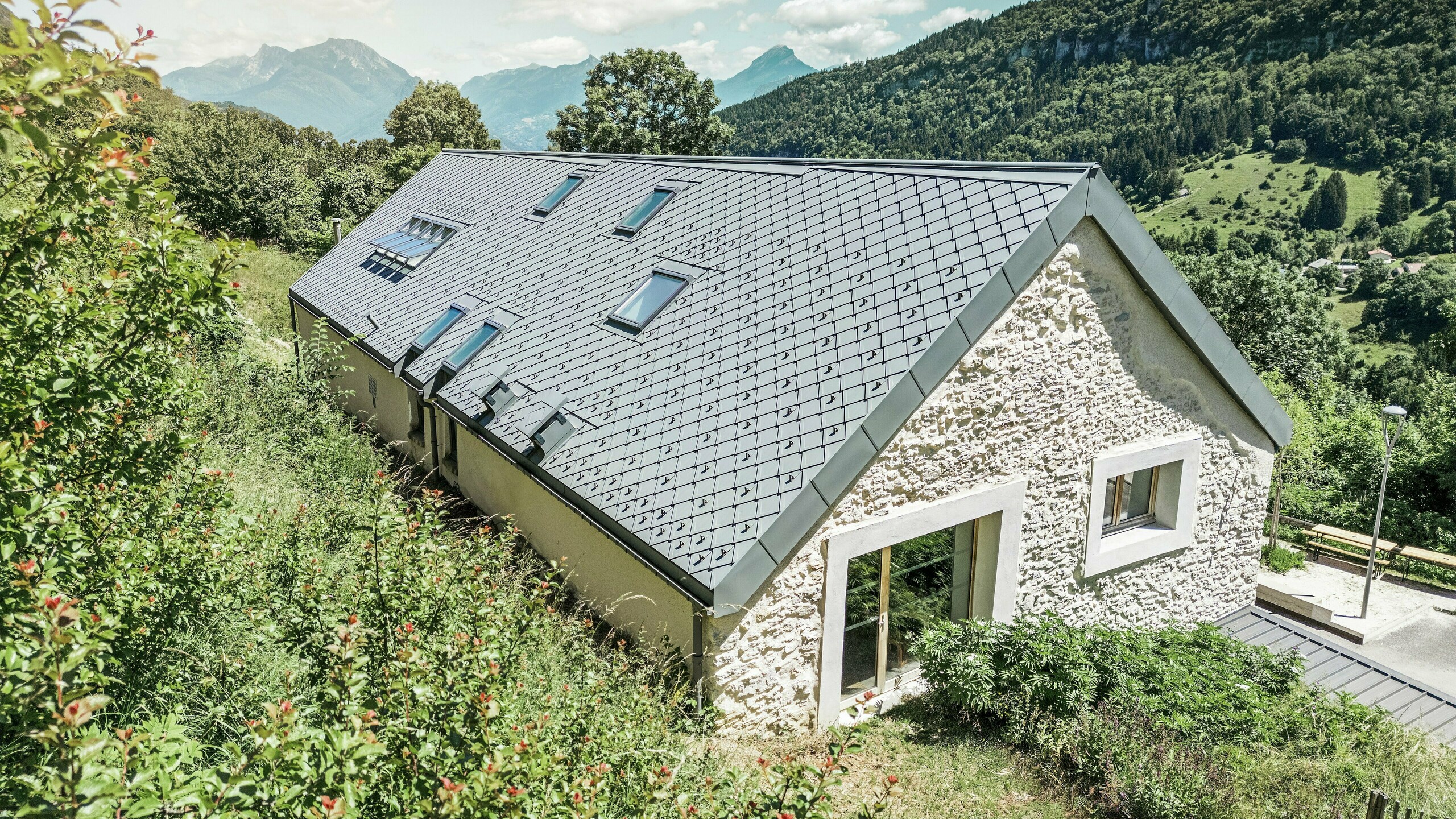 Vue du ciel de la ferme rénovée "La Grange à Gaby" à Engins, France. Le bâtiment est recouvert de losanges de toiture PREFA 29 × 29 durables, qui combinent une protection moderne avec une esthétique traditionnelle. Le paysage de montagne idyllique souligne l'intégration harmonieuse dans la nature.