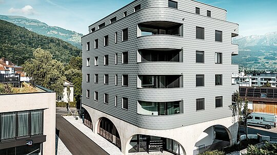 Vue extérieure de la Bernerhaus à Buchs, caractérisée par sa forme triangulaire audacieuse et ses balcons aux courbes douces. La façade en bardeau gris souris PREFA P.10 enveloppe le bâtiment comme une peau d'aluminium à écailles, créant une façade à la fois précise et dynamique. Légère et résistante aux intempéries, la façade en aluminium PREFA se distingue particulièrement au-dessus du socle en béton massif et de ses élégantes arcades. Durable, nécessitant peu d'entretien et d'une grande stabilité dimensionnelle, elle affirme une architecture moderne sur fond de montagnes alpines.