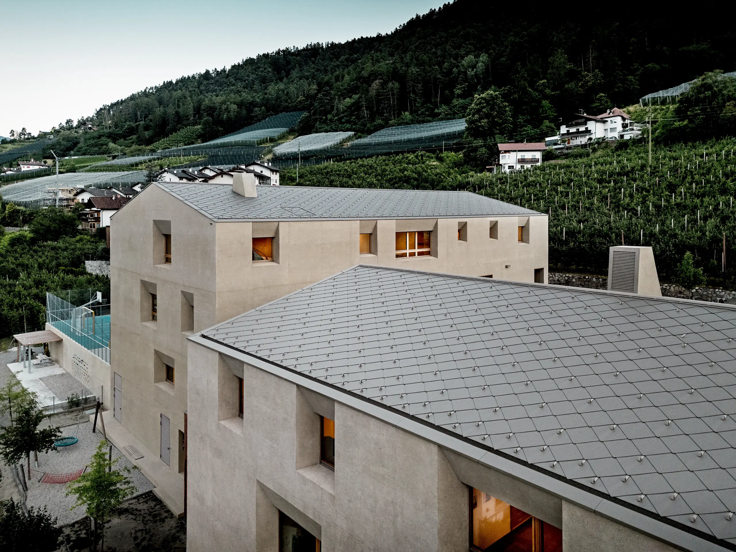 Vue du ciel, le centre éducatif de Riffian, dans le Tyrol du Sud, révèle ses remarquables losanges de toiture PREFA 44 × 44, déclinés dans le coloris Bronze P.10. Ce revêtement en aluminium, d’une grande précision, se déploie avec sérénité sur le jardin d’enfants et l’école primaire ; offrant une protection durable et résistante aux intempéries, il intègre de manière spectaculaire cette architecture moderne au cœur des vergers et du versant montagneux.