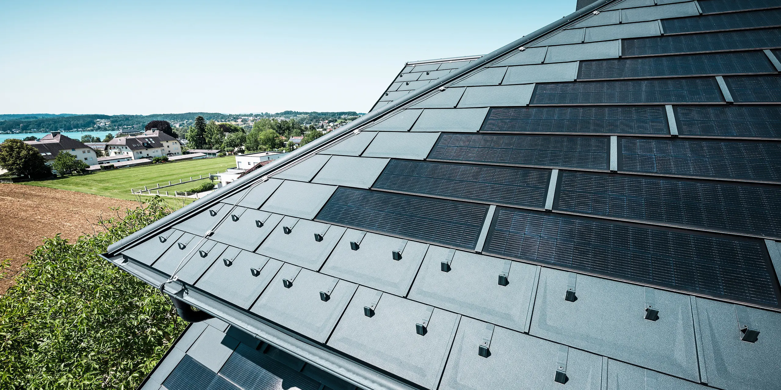 [Translate to Français:] Nahaufnahme des neu sanierten Daches der Zenz'n Stub'n in Schörfling am Attersee, eingedeckt mit langlebigen PREFA Dachplatten R.16 und innovativen PREFA Solardachplatten in der Farbe P.10 Anthrazit. Das Aluminiumdach verbindet modernes Design mit nachhaltiger Energiegewinnung. Die robuste Dachentwässerung in passendem P.10 Anthrazit fügt sich harmonisch in das Gesamtbild ein. In der Ferne ist im Hintergrund der blaue Attersee zu erkennen.