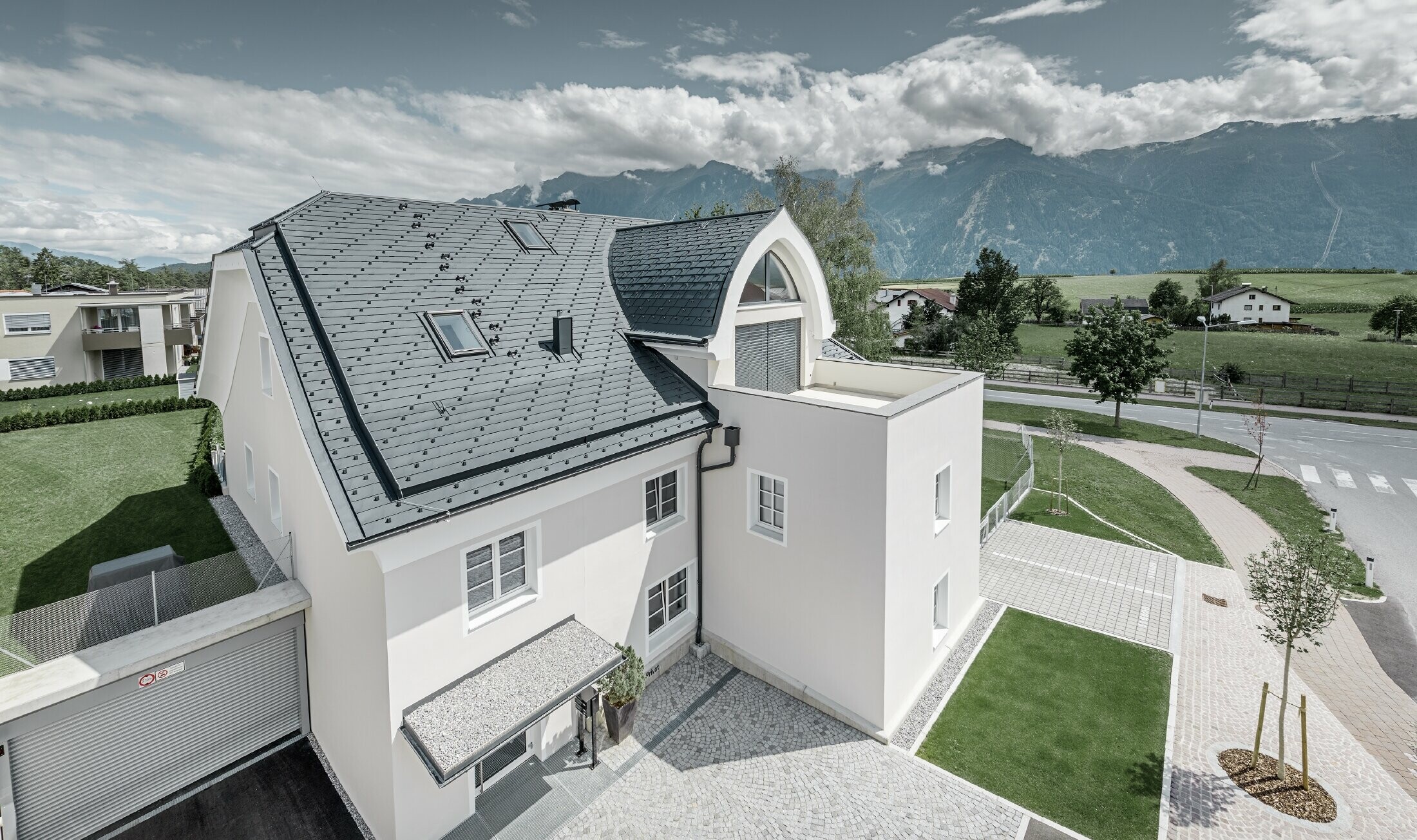 Bâtiment d’habitation rénové avec une nouvelle toiture PREFA ; le toit à demi croupe avec lucarne en ogive et loggia a été recouvert de bardeaux en alu de PREFA couleur anthracite. Des arrêts neige, des encadrements de fenêtre et des gouttières de PREFA ont également été employés.