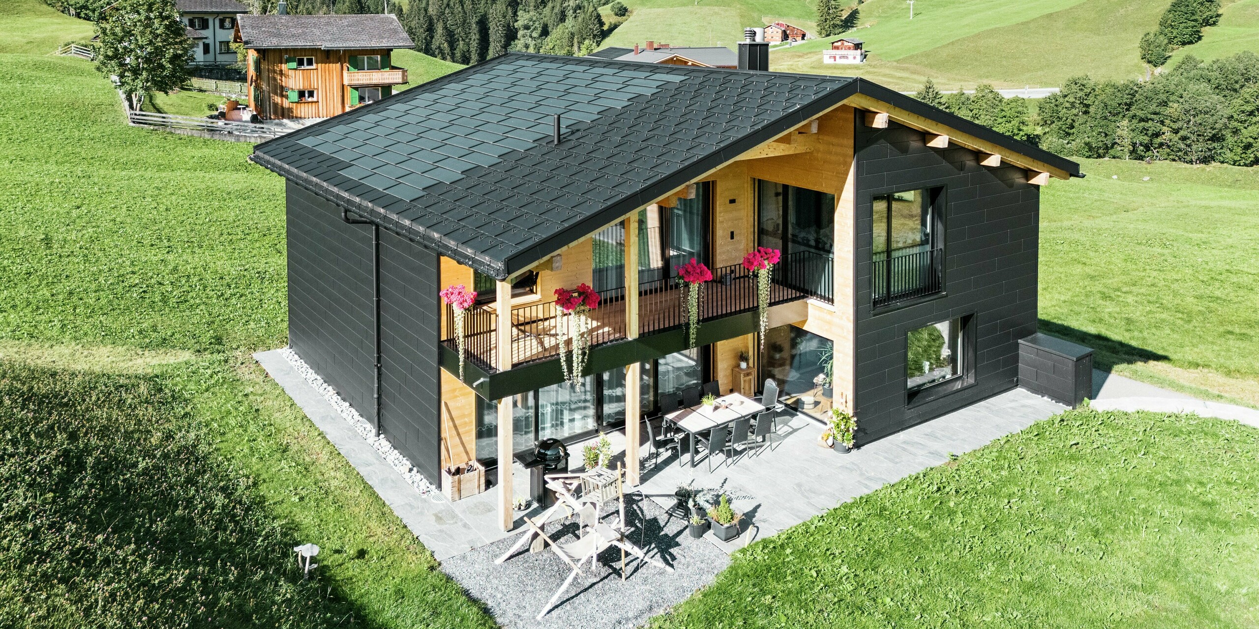 Cette élégante maison individuelle située dans les environs vallonnés d'Ascharina (Suisse) montre comment l'aluminium et le bois peuvent être parfaitement combinés. Les robustes panneaux de façade PREFA Siding.X en noir P.10 s'harmonisent avec le toit composé de panneaux solaires PREFA et de panneaux de toiture R.16 dans le même revêtement de couleur. L'enveloppe en aluminium durable protège le bâtiment de manière fiable contre les défis du climat alpin.