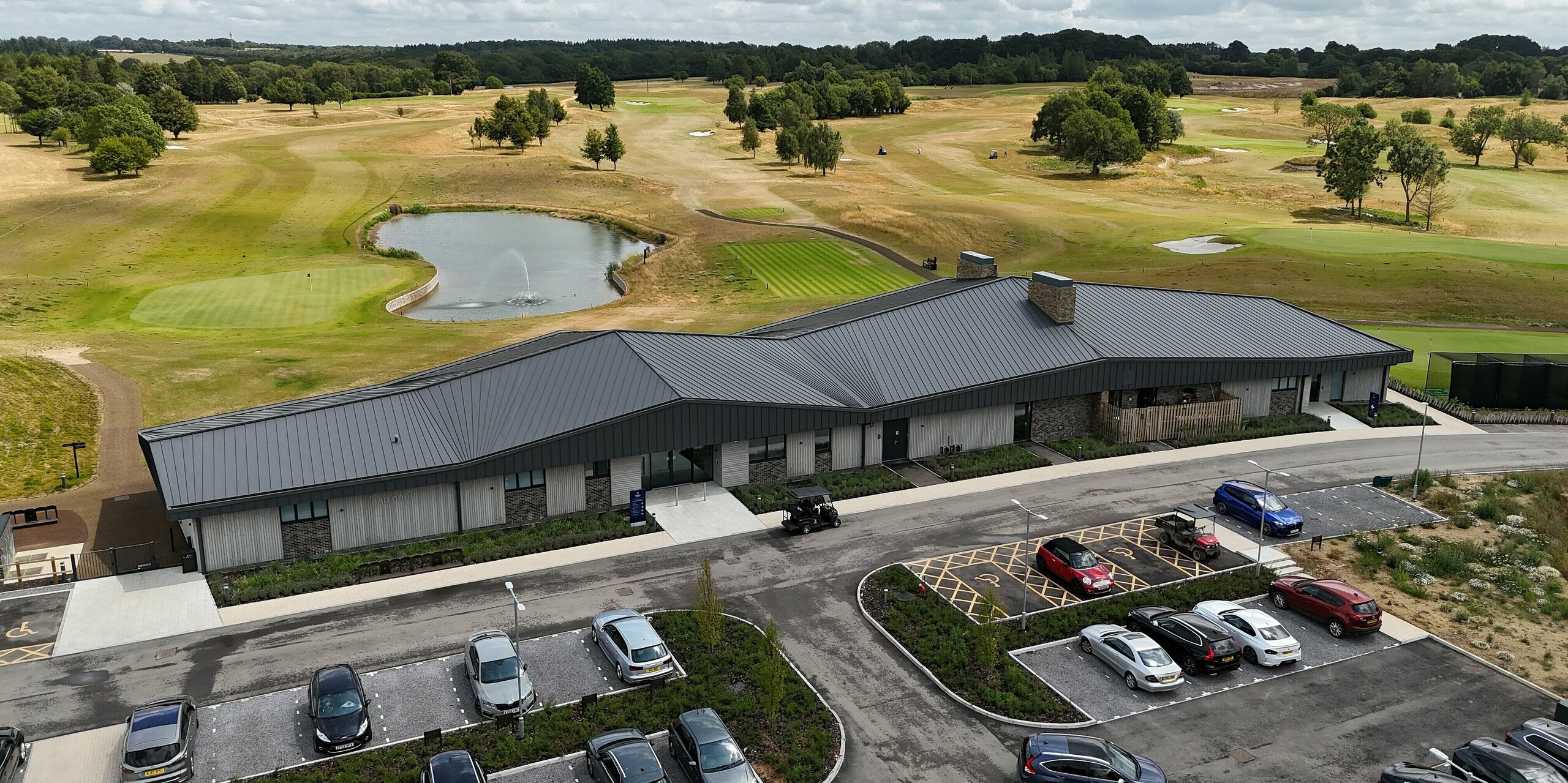Le nouveau bâtiment moderne du Basingstoke Golf Club, dans le Hampshire, impressionne par son élégante toiture à joints debout PREFALZ, de couleur gris sombre P.10. La forme dynamique du toit et la façade raffinée en bois et en pierre s'intègrent harmonieusement au vaste paysage du golf. Le bâtiment bénéficie d'une situation pittoresque au bord de l'eau, entouré d'espaces verts soignés. Au premier plan, on aperçoit le parking avec des véhicules et une voiturette de golf. L'aluminium durable et nécessitant peu d'entretien de la toiture offre une protection durable contre les intempéries et confère au club-house une esthétique contemporaine et fonctionnelle.
