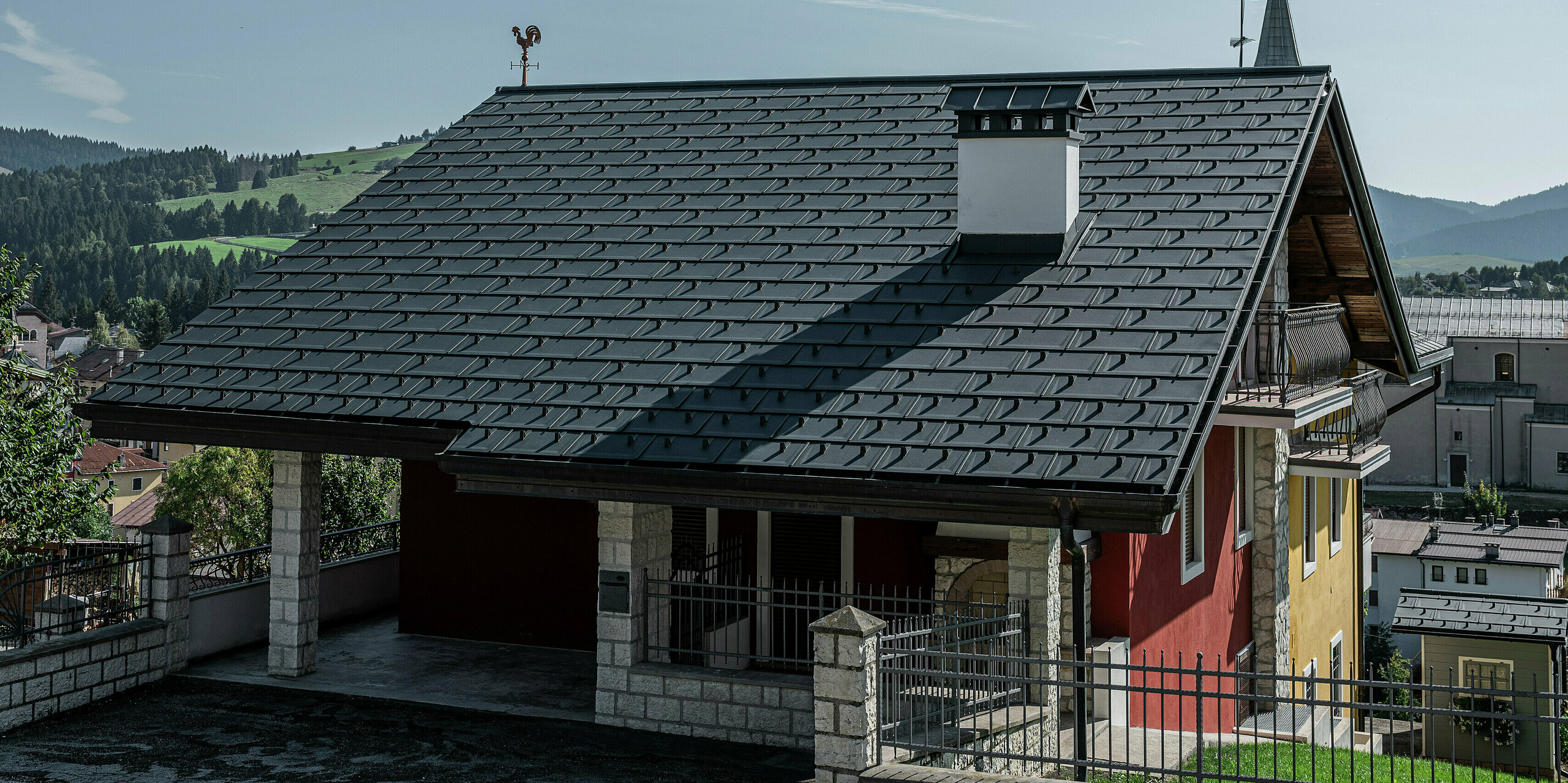 Vue détaillée de la zone d'entrée d'une maison individuelle à Gallio avec un toit PREFA robuste composé de R.16 en P.10 gris sombre. La couverture de toit en aluminium offre une apparence élégante et offre en même temps une protection maximale contre les éléments. La fabrication précise et le matériau durable garantissent une solution sans entretien pendant des décennies. La cheminée massive avec revêtement en pierre naturelle et la girouette traditionnelle ajoutent des accents élégants. Une combinaison parfaite de technologie moderne et de charme alpin.