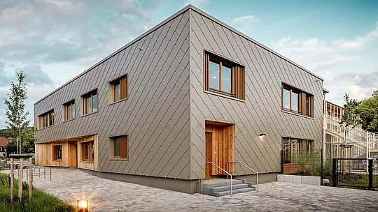La crèche moderne d'Alzenau séduit par ses lignes épurées et sa façade en aluminium PREFA couleur bronze de haute qualité. Composée de losange de façade PREFA de 44 x 44 cm, cette façade confère au bâtiment une allure élégante et structurée, unifiant visuellement les espaces fonctionnels en un ensemble harmonieux. Le bardage en bois aux entrées offre un contraste chaleureux et naturel avec la robustesse de l'aluminium. L'alliance d'une construction solide, d'éléments en bois et d'une façade ventilée à double paroi illustre parfaitement les pratiques de construction durable. L'enveloppe du bâtiment PREFA, nécessitant peu d'entretien, est non seulement recyclable, mais aussi extrêmement résistante aux intempéries – un atout majeur pour les établissements scolaires d'avant-garde répondant à des exigences architecturales élevées.