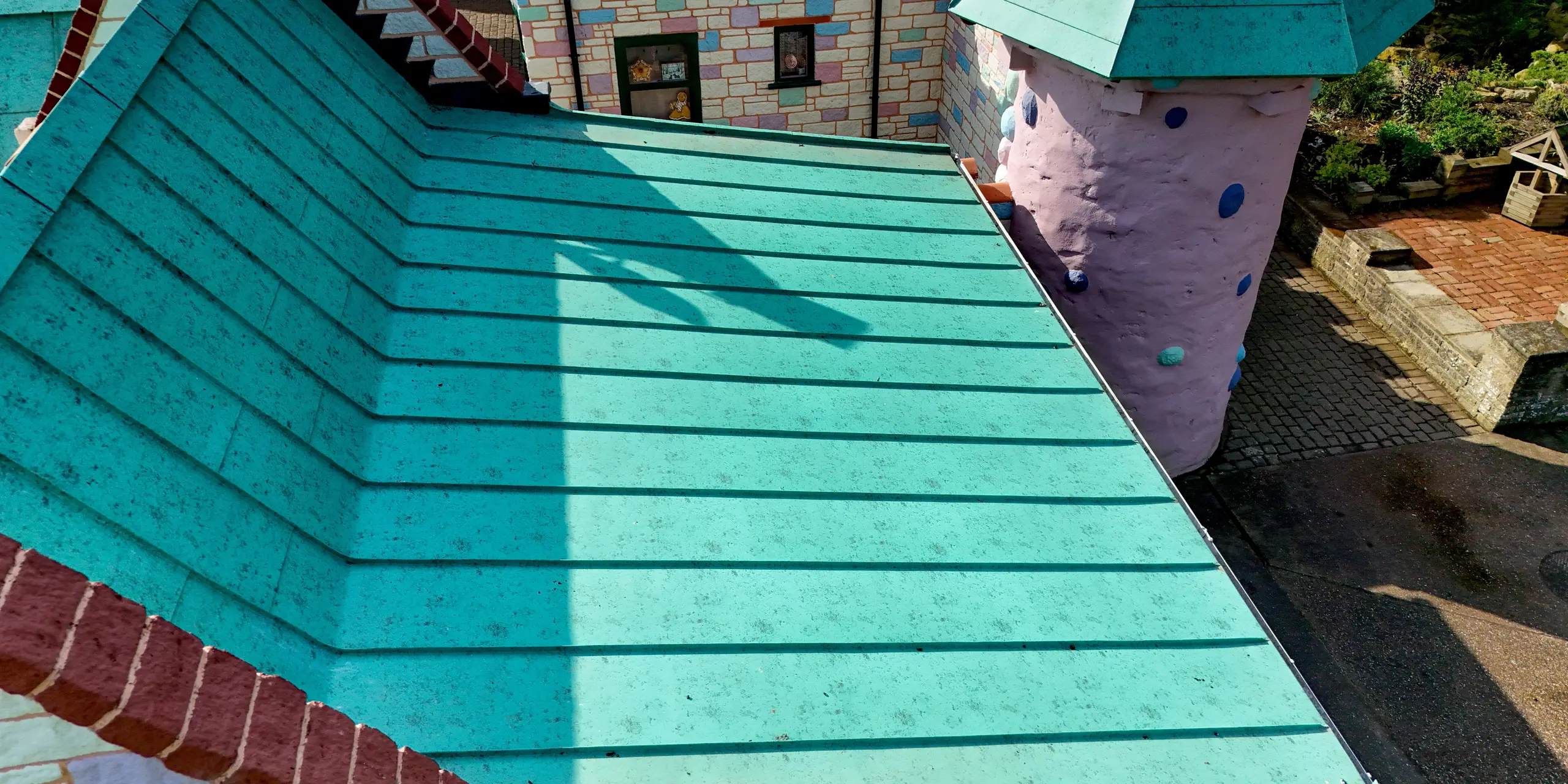 Gros plan sur une toiture à joints debout PREFALZ à Sundown Adventureland, mettant en valeur des lignes de joints parallèles et nettes qui créent une structure visuelle saisissante. La surface en aluminium P.10 vert de gris attire immédiatement le regard et souligne la précision moderne malgré l'architecture ludique du château. La finition impeccable des bords et des transitions témoigne d'une grande qualité d'exécution. La toiture PREFA impressionne non seulement par son esthétique, mais aussi par sa fonctionnalité : grande durabilité, résistance à la corrosion et protection fiable contre le vent et les intempéries.