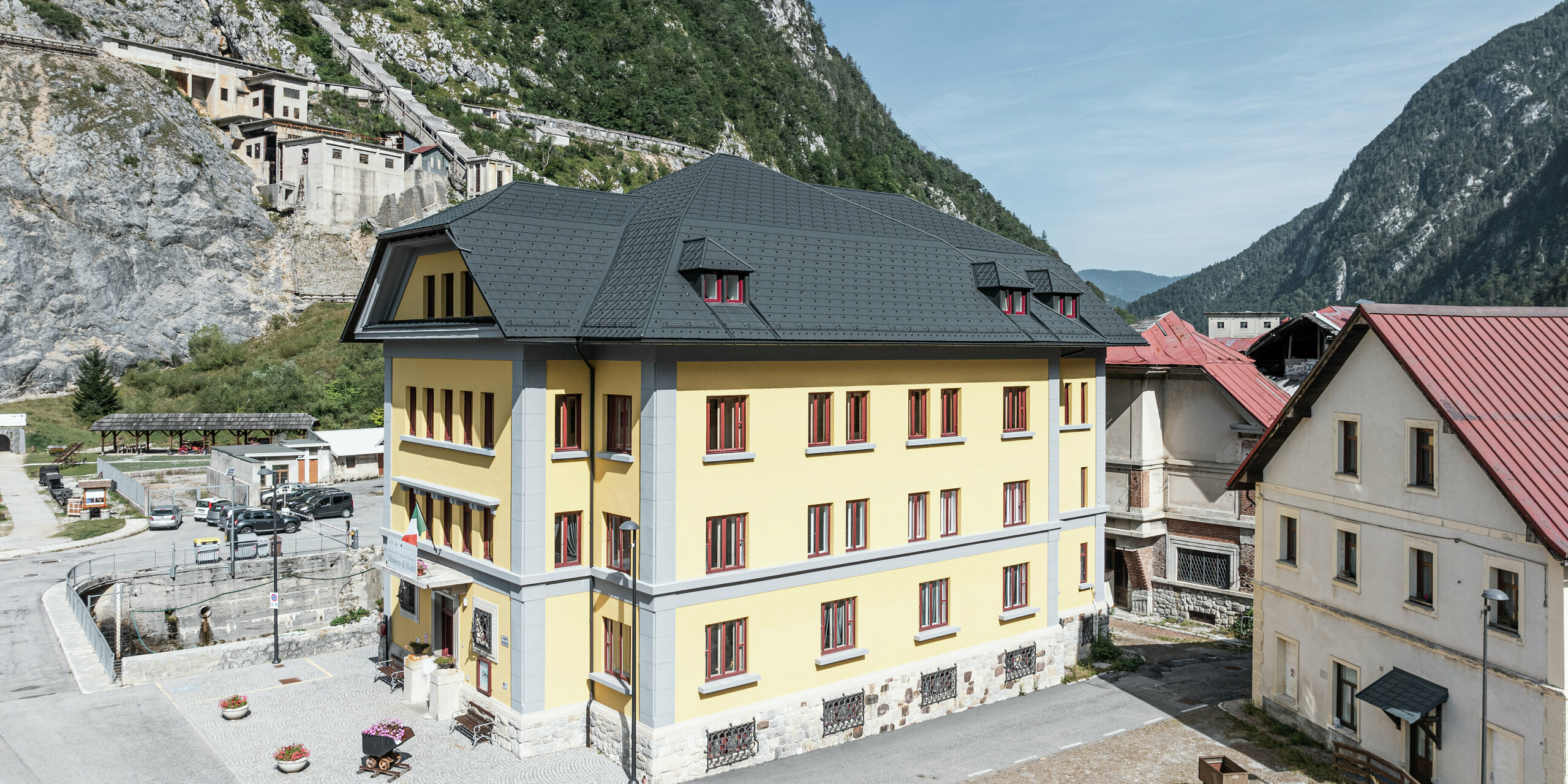 Bâtiment de musée historique Museo Cave del Predil en Italie avec façade jaune et toit en aluminium PREFA de couleur anthracite (tuiles P.10 anthracite). Paysages de montagne impressionnants et environnement alpin en arrière-plan. Le toit impressionne par sa durabilité et sa robustesse - idéal pour les conditions météorologiques alpines extrêmes.