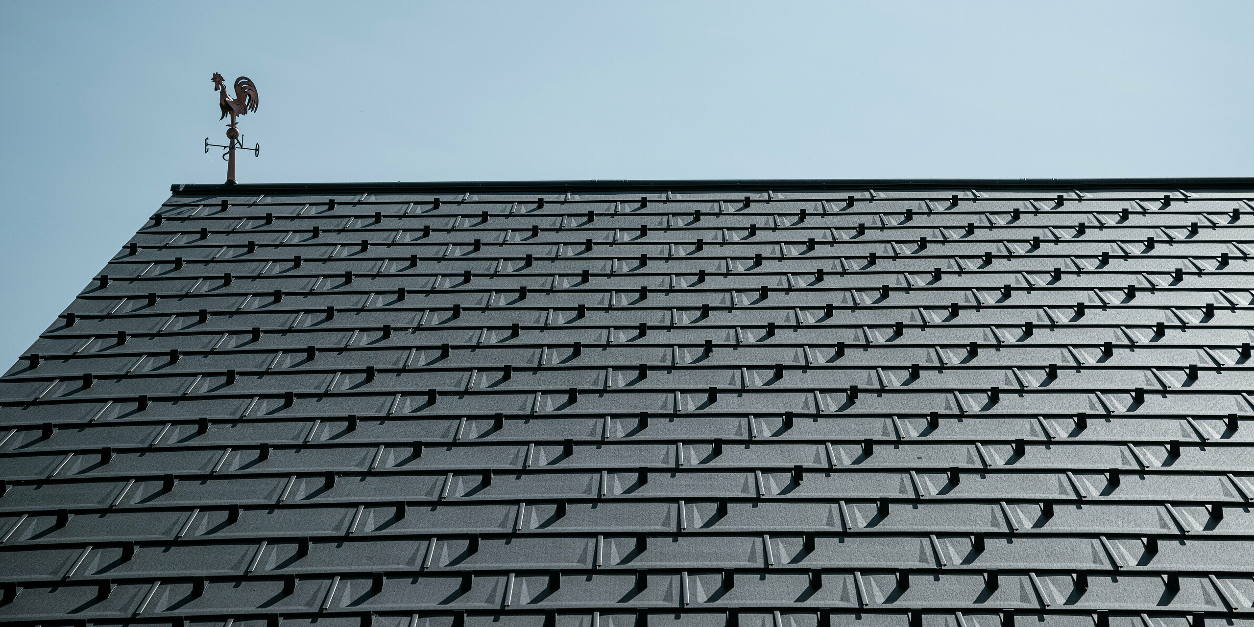 Vue détaillée d'une toiture PREFA avec les R.16 en P.10 gris sombre. L'aspect clair et moderne des panneaux de toiture en aluminium garantit une solution de toit durable, résistante aux intempéries et sans entretien. Les crochets à neige offrent une sécurité supplémentaire dans les régions enneigées. Le toit est couronné d'une girouette classique qui allie tradition et technologie moderne. Grâce aux R.16 robustes PREFA, le toit reste stable et esthétique pendant des décennies.