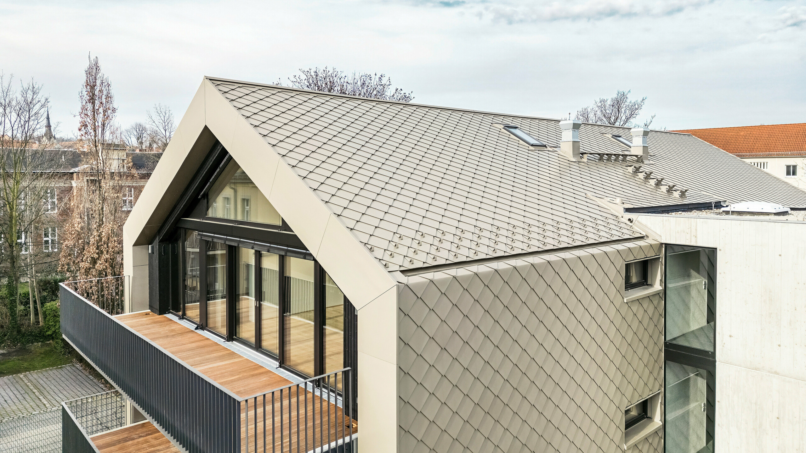 Vue plongeante d'un immeuble d'appartements moderne de la Chemnitzer Straße (Dresde) avec une enveloppe de bâtiment en losanges en aluminium PREFA 29 × 29 en bronze. L'image montre l'élégante charpente du toit, les grandes façades vitrées et les balcons ainsi que l'intégration harmonieuse du bâtiment dans l'environnement. Les matériaux de haute qualité soulignent l'architecture durable du bâtiment.