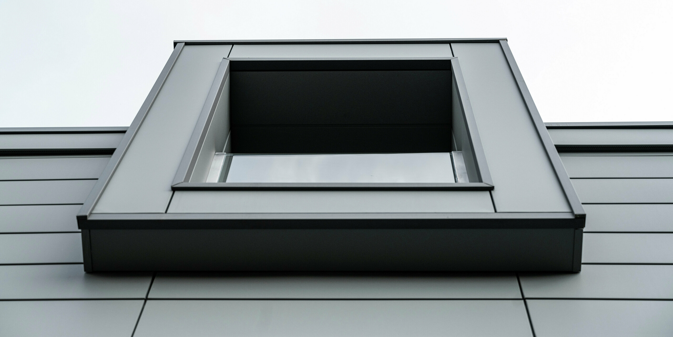 Vue rapprochée d'en bas d'une fenêtre en porte-à-faux d'un complexe résidentiel à Faloppio, en Italie. La forme claire et géométrique de la fenêtre, combinée à la façade en aluminium durable PREFA Siding, souligne l'architecture moderne et le design minimaliste du bâtiment.