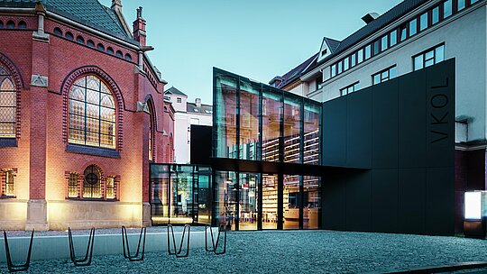 L'image met en valeur une réalisation architecturale remarquable à Olomouc, qui fusionne harmonieusement l'église rouge historique et une extension moderne. La façade noire du nouveau bâtiment, construite en panneaux composites en aluminium PREFABOND, contraste avec élégance avec les murs de briques rouges de l'église. De larges baies vitrées laissent entrevoir l'intérieur, dévoilant des étagères de livres et une atmosphère chaleureuse. La précision du choix des matériaux crée un équilibre harmonieux entre les éléments historiques et modernes. L'utilisation de matériaux durables et esthétiques comme PREFABOND garantit non seulement un style contemporain, mais aussi une qualité et une fonctionnalité pérennes. Ce projet architectural illustre avec brio comment l'ancien et le nouveau peuvent fusionner pour créer un ensemble urbain unique.