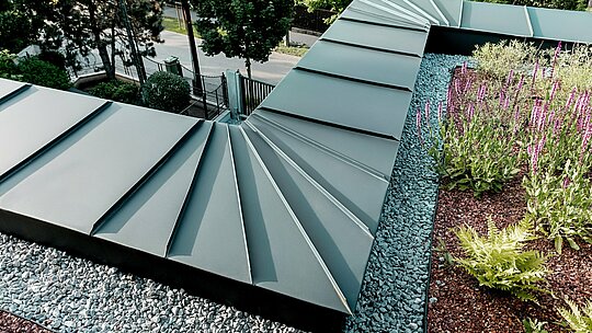 La photographie présente un gros plan détaillé du parapet de la toiture d'une villa multigénérationnelle à Budapest, revêtue d'aluminium PREFALZ couleur P.10 anthracite. La disposition en éventail des panneaux converge précisément au bord du bâtiment, témoignant de la grande formabilité et de la qualité de fabrication des produits en aluminium PREFA. Le métal anthracite offre un contraste élégant avec la toiture végétalisée, agrémentée d'un lit de gravier et de plantes fleuries. Cette scène met en valeur la durabilité, la résistance aux intempéries et la technicité de la solution PREFA.