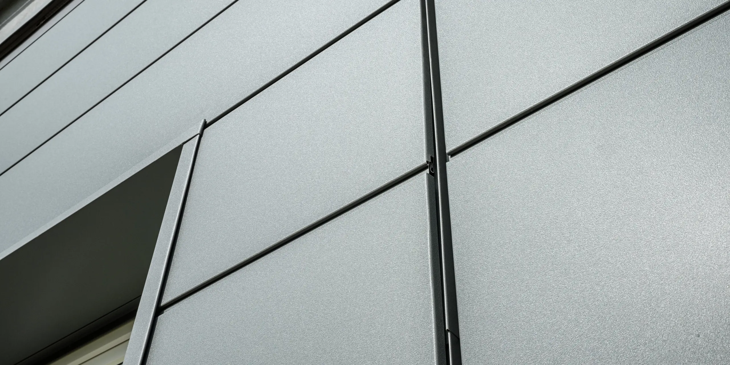La façade du bâtiment de la guilde des ramoneurs à Bindlach est revêtue d'un bardage moderne PREFA P.10 gris sombre. Les panneaux d'aluminium à la structure apparente sont non seulement esthétiques, mais aussi extrêmement résistants aux intempéries. Le gros plan révèle clairement la précision de la pose des panneaux métalliques et l'élégance minimaliste des lignes.