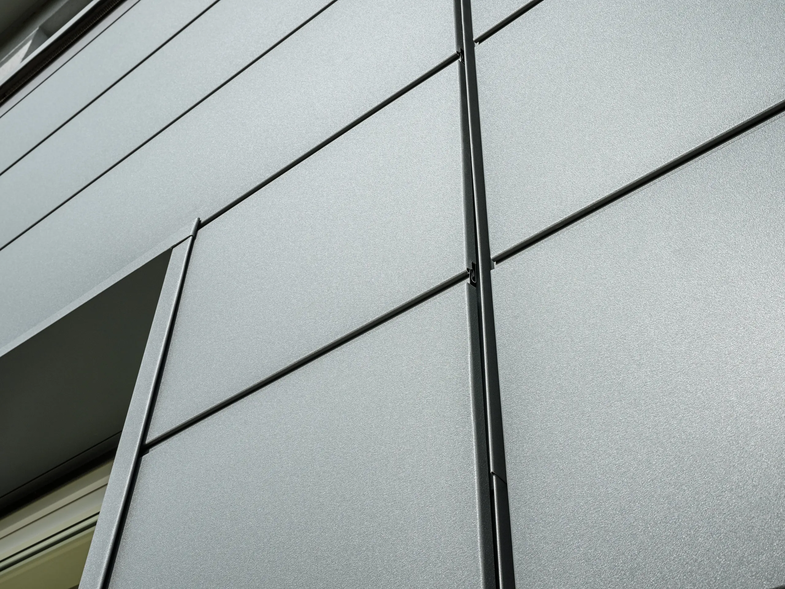 La façade du bâtiment de la guilde des ramoneurs à Bindlach est revêtue d'un bardage moderne PREFA P.10 gris sombre. Les panneaux d'aluminium à la structure apparente sont non seulement esthétiques, mais aussi extrêmement résistants aux intempéries. Le gros plan révèle clairement la précision de la pose des panneaux métalliques et l'élégance minimaliste des lignes.