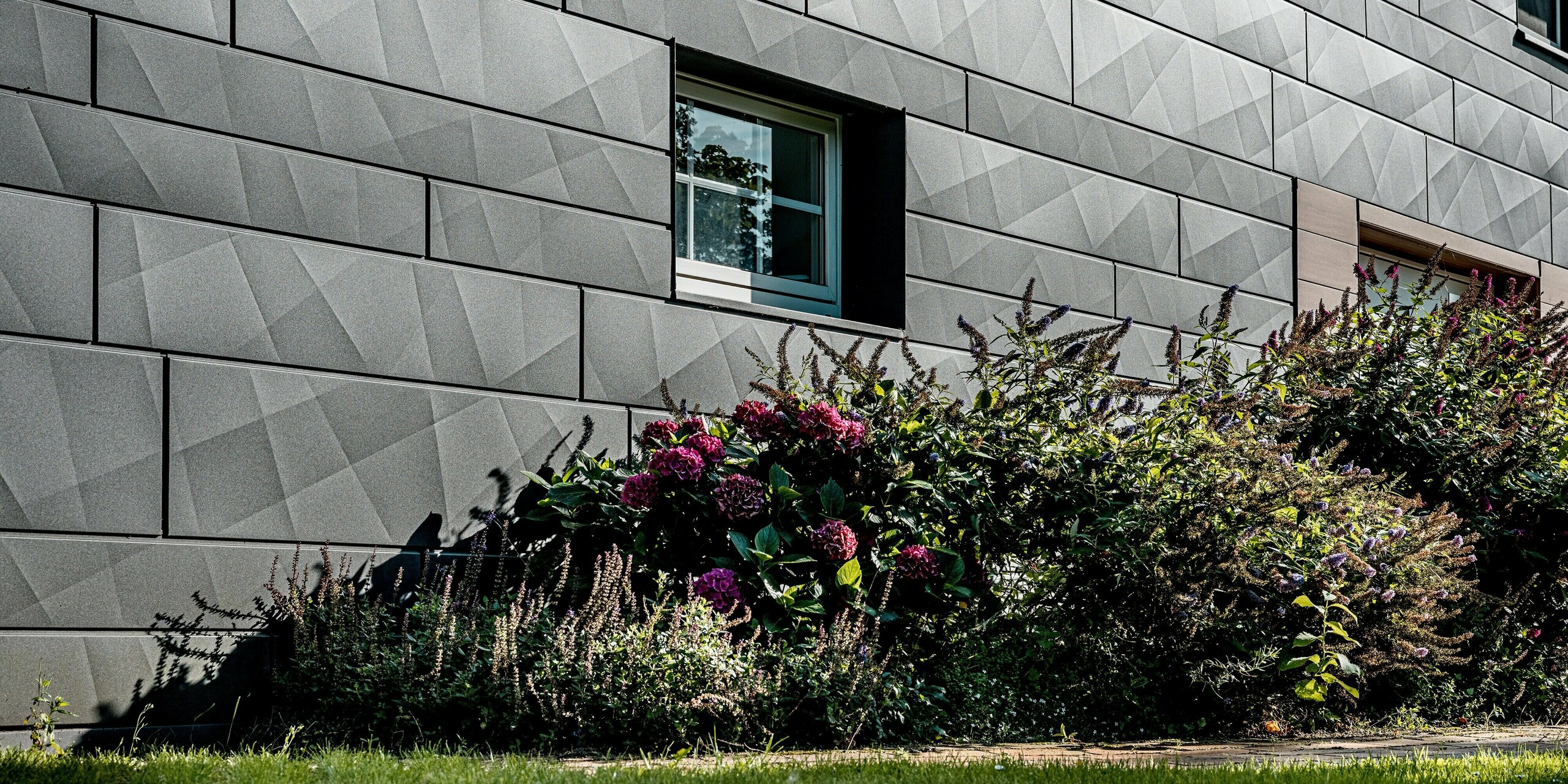 Nahaufnahme der sanierten Fassade eines Einfamilienhauses mit PREFA Siding.X in P.10 Dunkelgrau. Die großformatigen Aluminiumpaneele mit markanten Längs- und Querkantungen erzeugen eine lebendige, geometrische Oberflächenstruktur, die je nach Lichteinfall spannende Licht- und Schattenspiele zeigt. Ein präzise eingefasstes Fenster setzt einen klaren architektonischen Akzent und unterstreicht die hochwertige Verarbeitung. Im Vordergrund kontrastiert ein üppiges Blumenbeet mit farbigen Blüten und Grünpflanzen die kühle Eleganz der Aluminiumfassade. Die witterungsbeständigen, farbstabilen und pflegeleichten PREFA Aluminiumpaneele verbinden ausdrucksstarkes Design mit langlebiger Qualität und sind ideal für moderne, nachhaltige Fassadensanierungen.