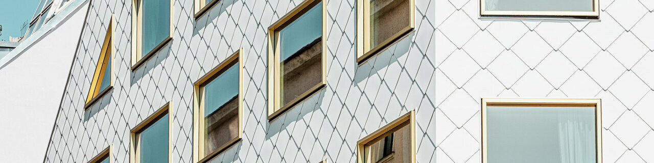 Un hôtel quatre étoiles moderne à Vienne, habillé de losanges de toit et de murs PREFA 44 × 44 en P.10 blanc pur. La façade présente une structure en forme de bloc avec une multitude de fenêtres à vitrage miroir, disposées selon un quadrillage net qui reflète les mouvements de l'environnement.