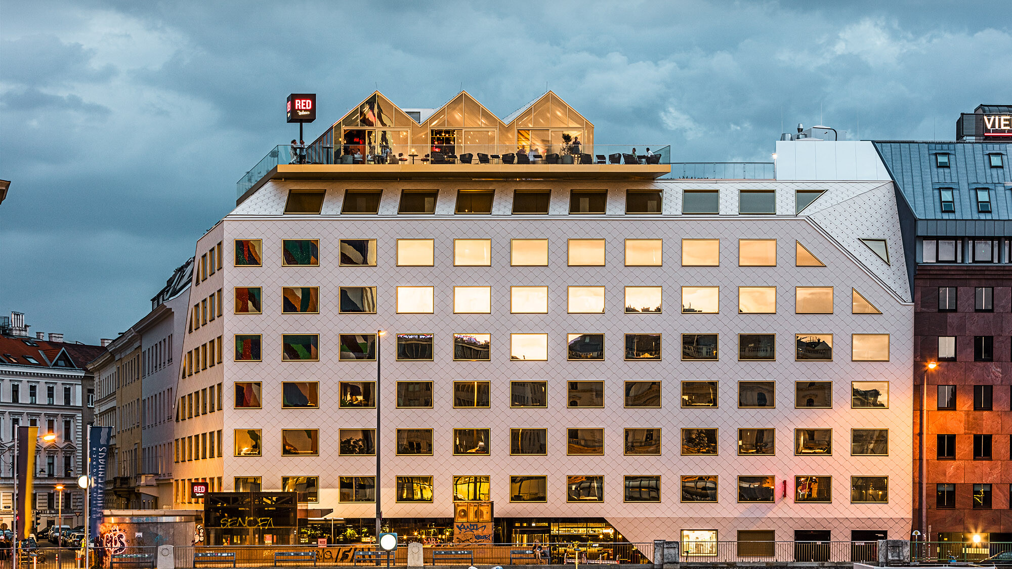 Une photo prise le soir de l'hôtel THE ROCK Radisson RED Vienna et de ses environs animés, les bâtiments d'en face se reflétant dans ses fenêtres.