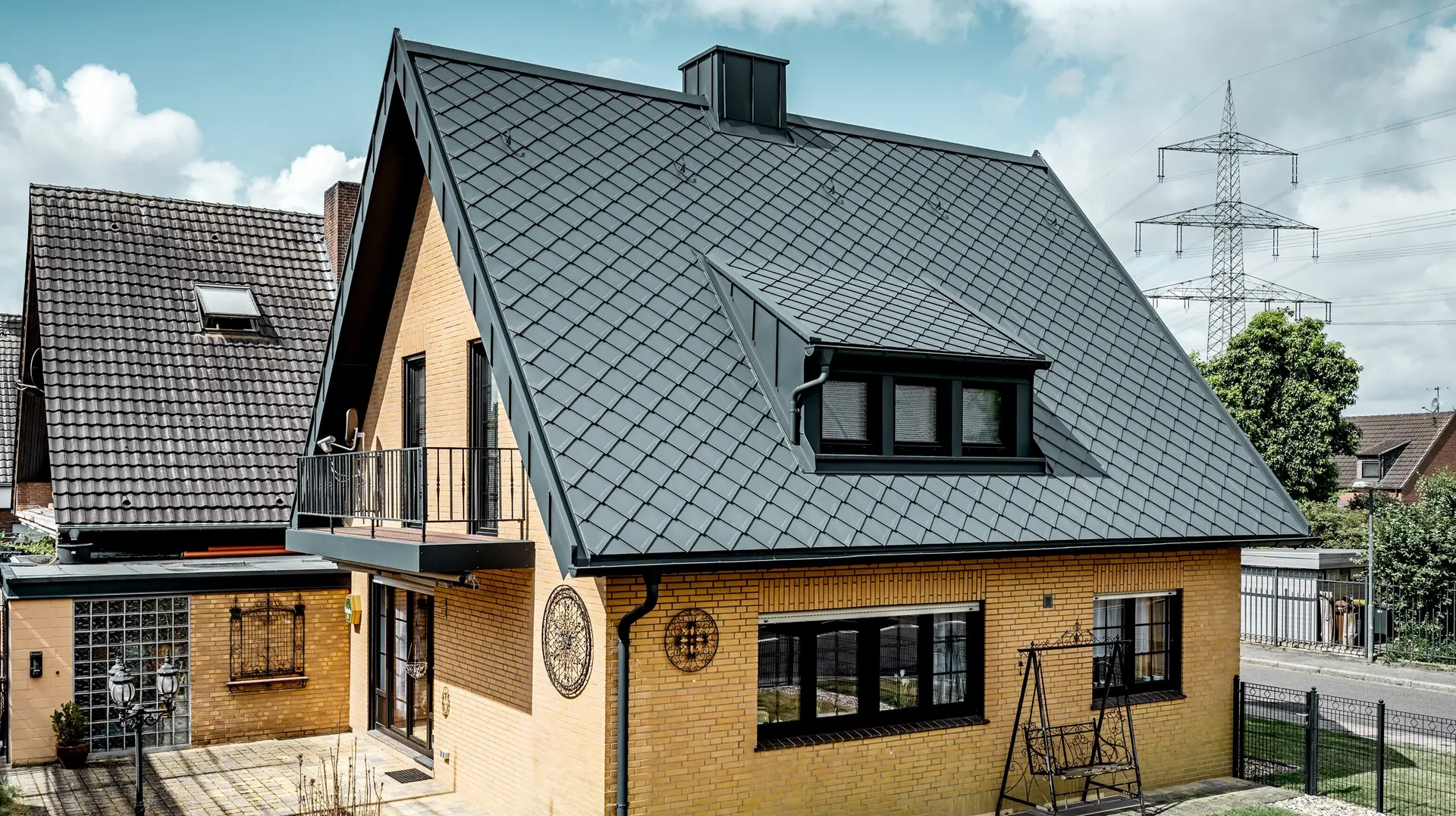 Maison individuelle en briques jaunes après la rénovation du toit avec un losange de toiture PREFA 29 × 29 en gris sombre ; toit en pente raide nouvellement recouvert avec un motif en losange, une lucarne et une cheminée intégrées, terrasse et jardin au premier plan.