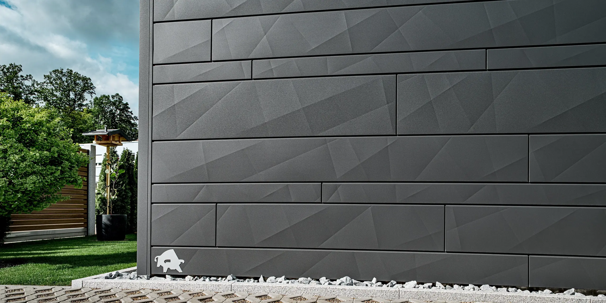 Vue détaillée de la façade en aluminium Siding.X PREFA, coloris P.10 gris sombre, sur une maison individuelle moderne à Heidenheim. Les panneaux aux lignes nettes, de largeurs variables et aux arêtes marquées, créent un motif géométrique distinctif qui produit de magnifiques jeux d'ombre et de lumière selon l'angle d'éclairage. Au premier plan, on aperçoit des pierres décoratives claires et un jardin soigné, tandis qu'à l'arrière-plan, se dessinent des arbres et une structure en bois. La surface en aluminium, résistante et durable face aux intempéries, souligne la qualité et la modernité de la façade. Le logo PREFA, le taureau, est visible sur le bord gauche de la façade.