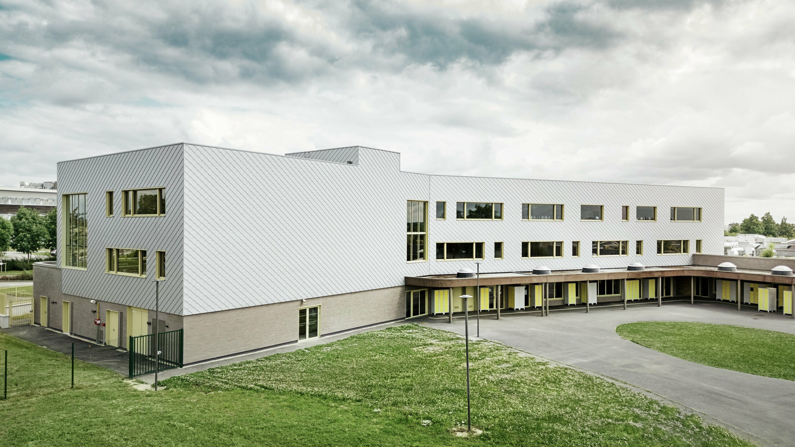 L'image montre l'arrière du collège Léontine Dolivet. Au premier plan, une vaste cour d'école pavée aux lignes épurées offre des espaces ouverts propices aux récréations et aux loisirs. Quelques espaces verts adoucissent l'environnement. À l'arrière-plan se dresse le bâtiment scolaire moderne, avec ses losanges de façade PREFA argent métallisé du plus bel effet, qui reflètent la lumière et créent un dynamisme certain. Plusieurs grandes fenêtres laissent entrer une abondante lumière naturelle. La scène a été photographiée en plein jour, sous un ciel légèrement nuageux, ce qui met particulièrement en valeur la surface métallique.