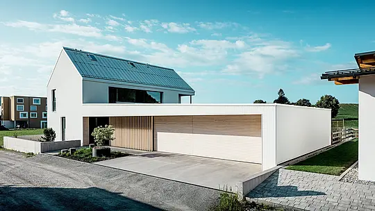 [Translate to Français:] Das Bild zeigt das moderne Einfamilienhaus in Freistadt, das sich durch seine schlichte, geometrische Form und die Kombination von reinweißer PREFALZ-Aluminium-Fassade und Holzvertäfelungen auszeichnet. Die Holzverkleidung am Garagentor bietet einen warmen, natürlichen Kontrast und fügt sich perfekt in das moderne Design ein. Das Dach, ebenfalls mit PREFALZ gedeckt, sorgt für eine nahtlose, wetterfeste Oberfläche. Diese Kombination aus pflegeleichtem Aluminium und Holz schafft eine ästhetisch ansprechende und funktionale Lösung für nachhaltige Architektur. Der weitläufige, grüne Außenbereich und der klare Himmel im Hintergrund unterstreichen die moderne Lebensqualität, die dieses Haus bietet.