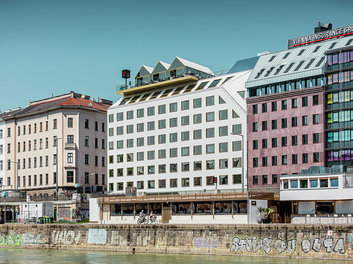 Vue sur les rives du canal du Danube à Vienne, avec une série de bâtiments d'architecture différente. Au premier plan, l'hôtel "THE ROCK Radisson RED Vienna", reconnaissable à sa façade blanche caractéristique en aluminium PREFA avec des losanges de toit et de façade 44 × 44 en P.10 blanc pur.