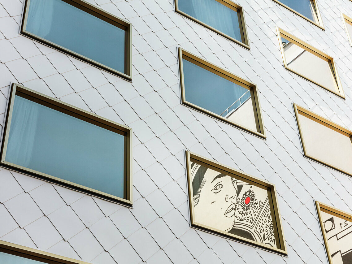 Détail de la façade de l'hôtel "THE ROCK Radisson RED Vienna", habillée de losanges de toit et de murs PREFA 44 × 44 en P.10 blanc pur. Parmi les nombreuses fenêtres à vitrage miroir et aux cadres dorés, une fenêtre se distingue par une œuvre d'art encastrée, représentant le portrait d'une personne avec un accent rouge prononcé