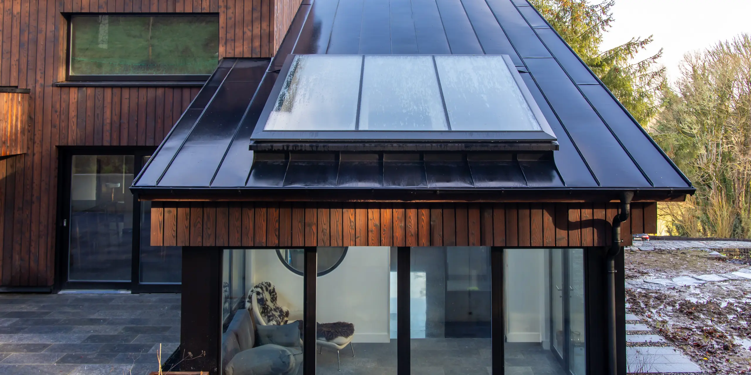 Détail de la véranda de la Maison Holywell à Sligo : vue de face, le regard est immédiatement attiré par la toiture en aluminium PREFALZ P.10 Noir, à l’exécution impeccable, qui épouse avec élégance la forme compacte du toit. Les joints debout nets structurent la surface et encadrent la large verrière. La surface lisse reflète la lumière et souligne la qualité de la réalisation. L’aluminium PREFA est reconnu pour sa durabilité, sa résistance à la corrosion et sa protection fiable contre les intempéries.