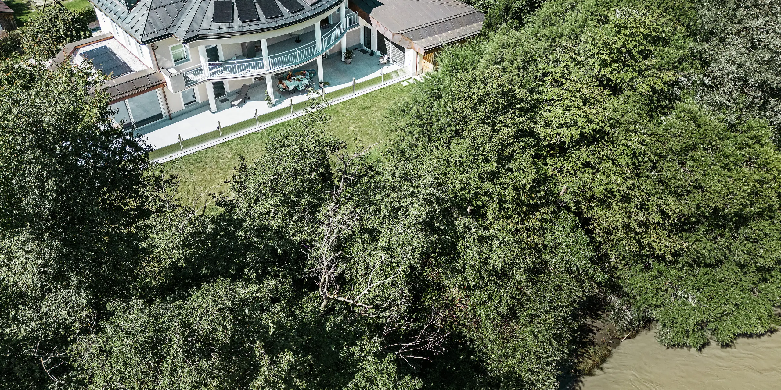Photographie aérienne d'une maison individuelle moderne à Schwaz, dans le Tyrol, située directement au bord d'une rivière. Le système de protection contre les crues PREFA installé le long de la terrasse, avec ses batardeaux en aluminium, forme une barrière sécurisée contre le cours d'eau adjacent. La perspective surélevée souligne la proximité immédiate de la maison avec le cours d'eau et l'importance d'une protection efficace du paysage et de la propriété.