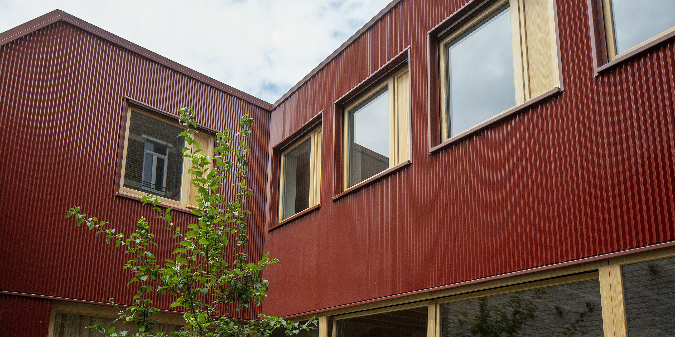 La cour intérieure rouge de Lille impressionne par le jeu dynamique des profils verticaux des poutres PREFA, du bois chaleureux et des grandes fenêtres. La couleur rouge oxyde intense crée un contraste harmonieux avec le vert frais des plantes et constitue un élément décoratif durable et sans entretien, à fort impact architectural.